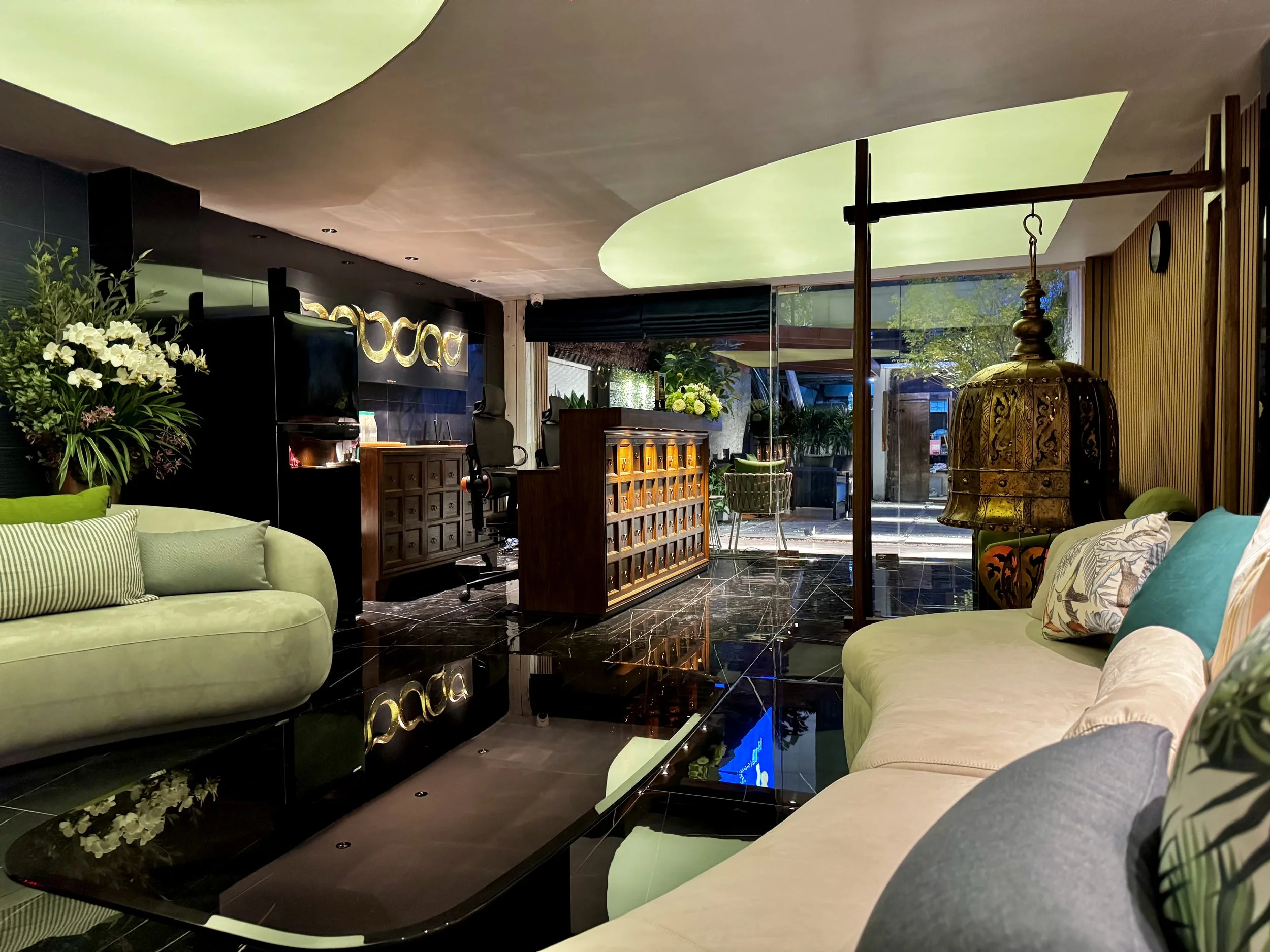 Modern hotel lobby with green and beige sofas, decorative pillows, a black coffee table, large floral arrangement, ornate hanging lantern, reception desk, office chair, and view of outdoors through glass windows.