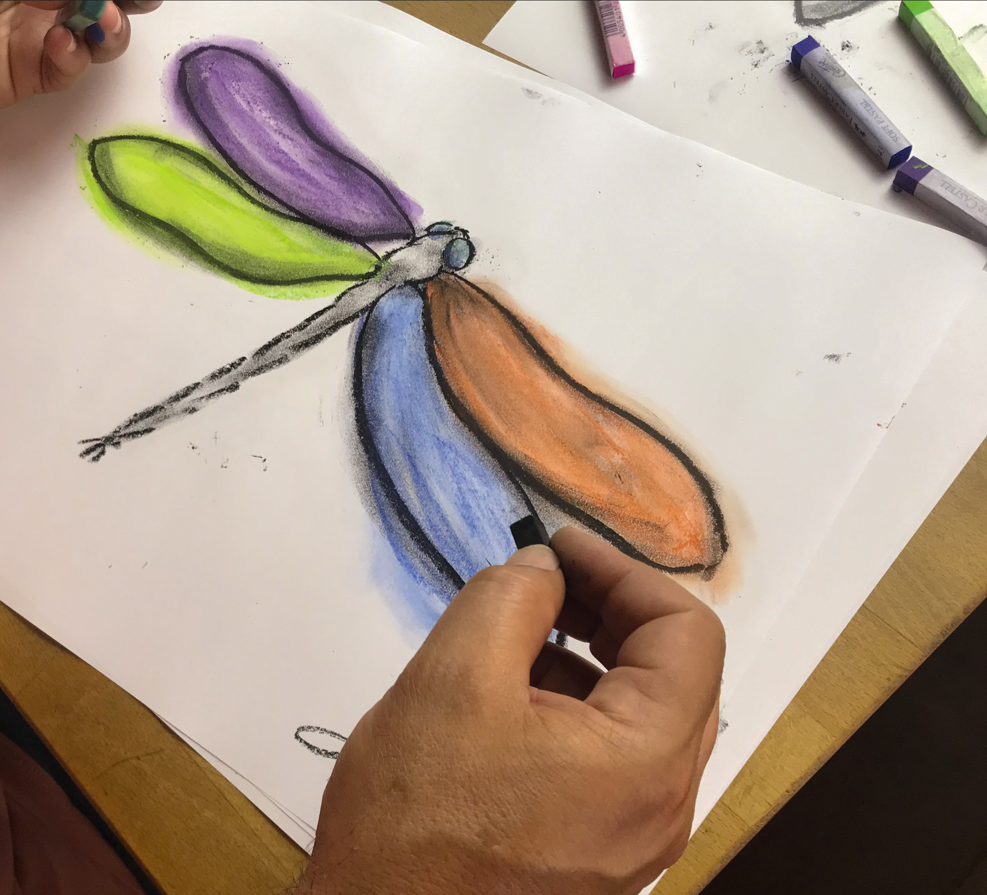 Desenho de uma libélula colorida feita com giz de cera, com asas verdes, roxas e alaranjadas, e corpo cinza, azul e preto, pintada sobre uma folha de papel branco.
