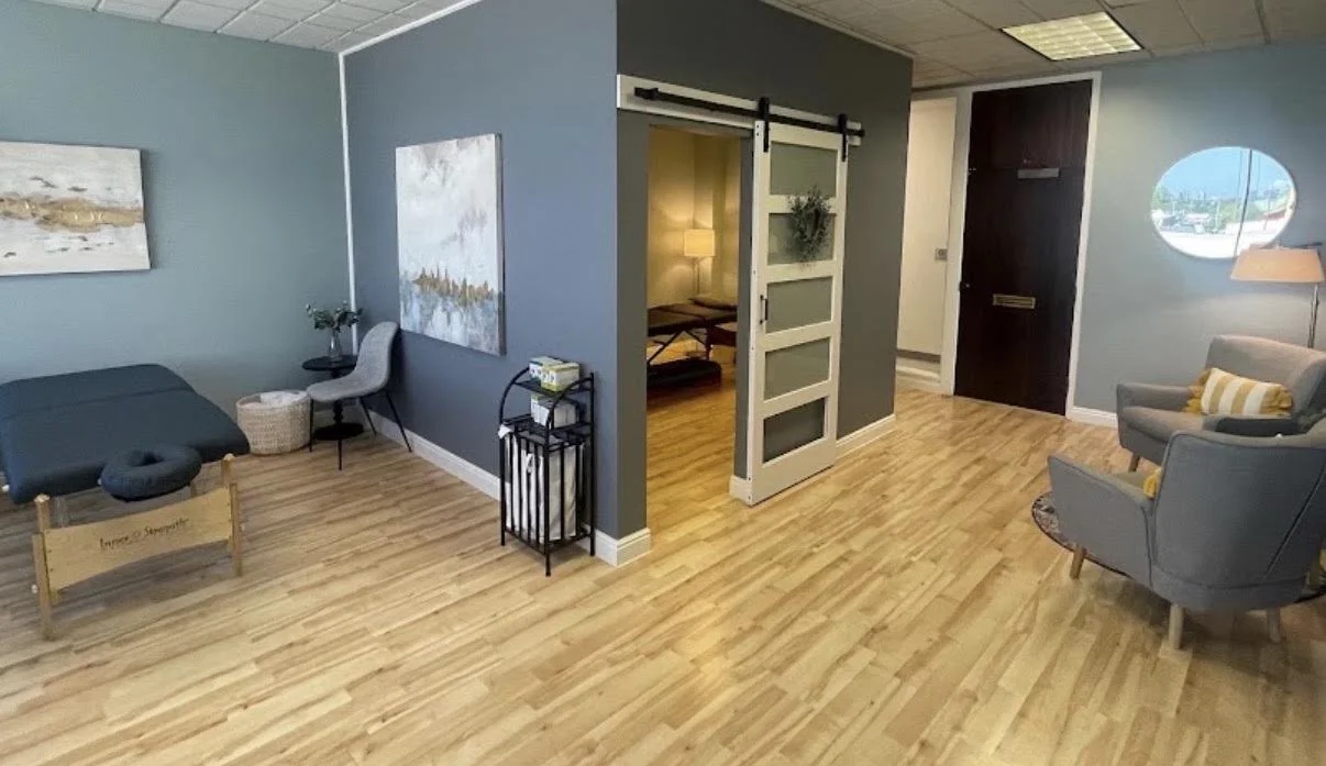 A room with blue walls and wooden flooring, furnished with a massage table, a chair, a small table with a plant, a basket, a black stand with tissues, a gray armchair with a yellow-striped pillow, a circular window, a doorway with a sliding barn door, and a view of a hallway with a close door and standing lamp.