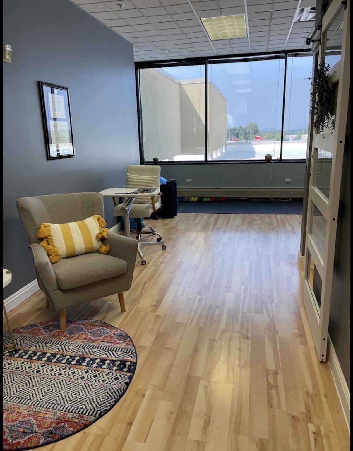 An office space with wooden flooring, a grey armchair with a yellow and white striped pillow, a small round rug, a beige chair at a desk, and large windows showing a parking lot outside.