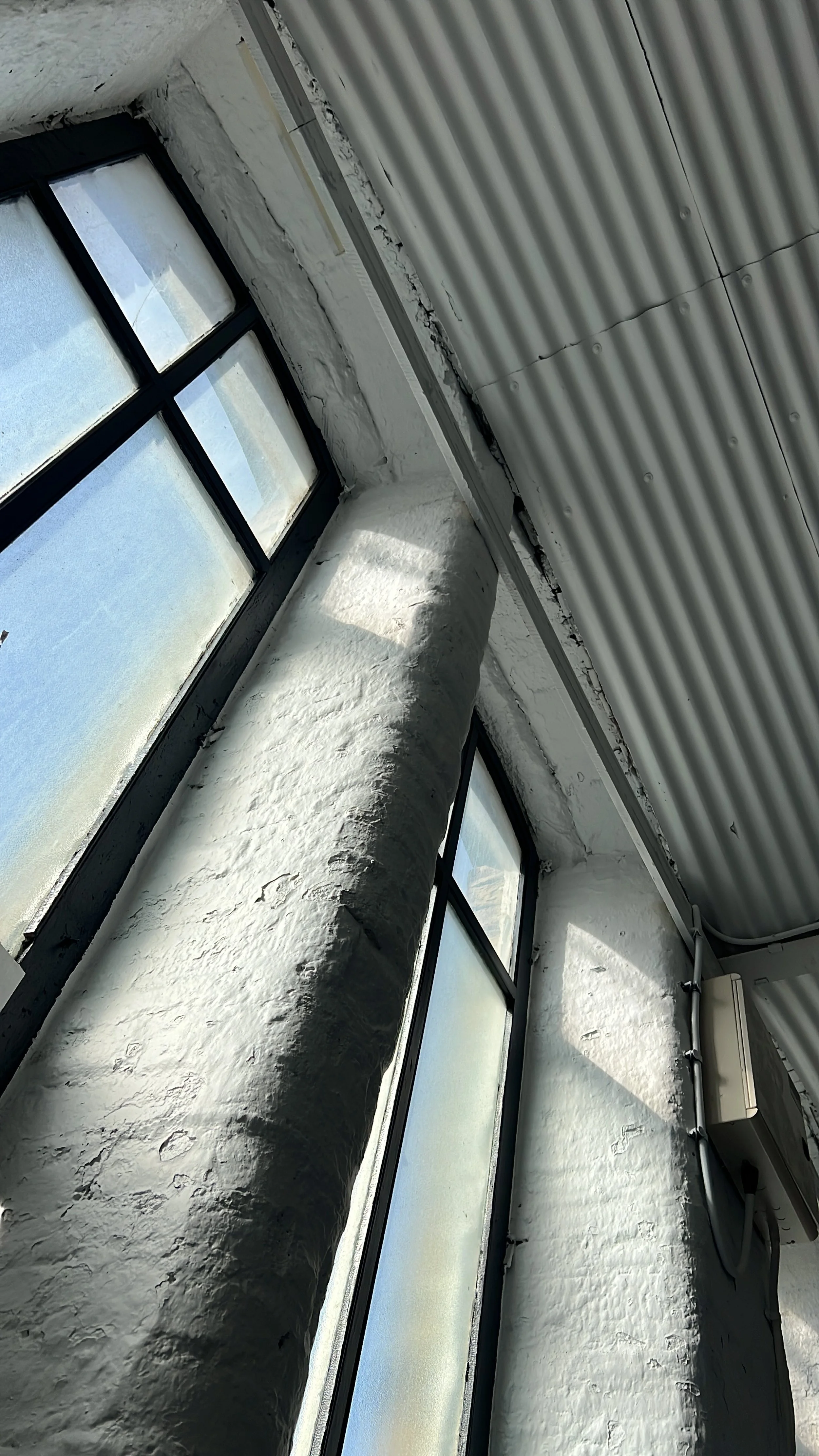 Interior of a building with large industrial-style windows, white painted textured brick walls, and an exposed corrugated metal ceiling.