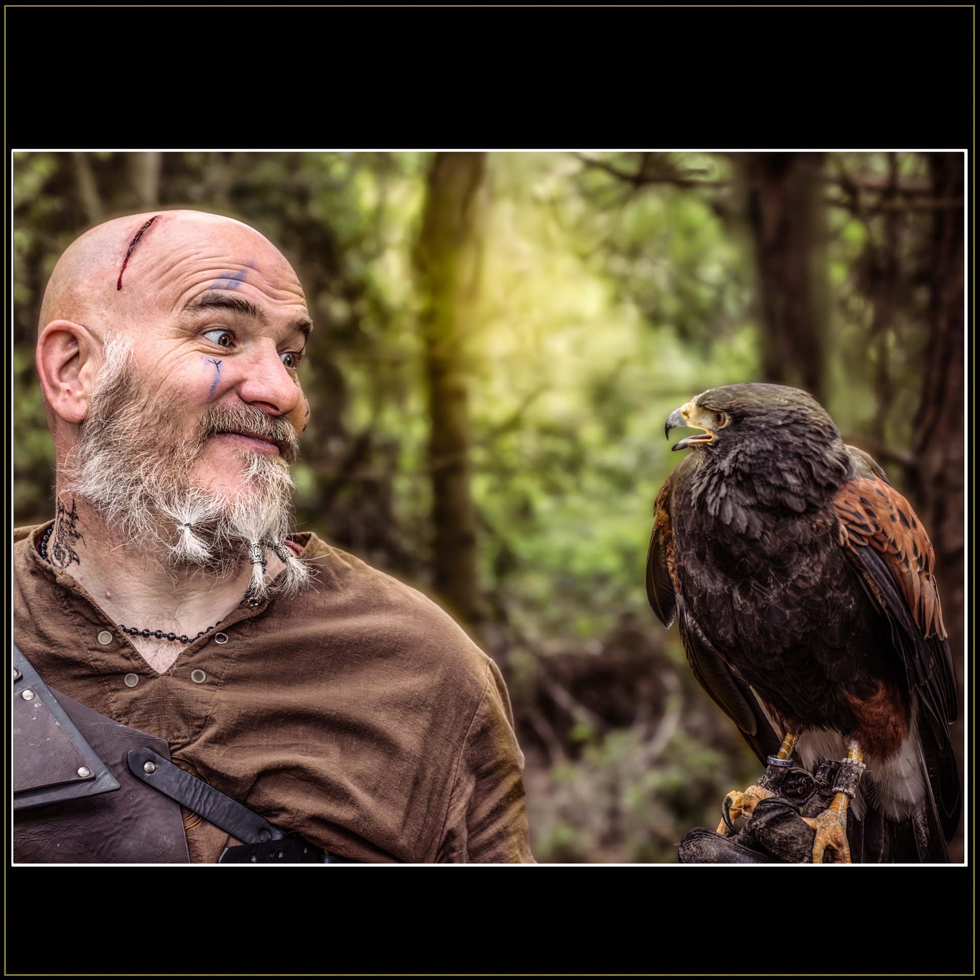 Don’t Chirp at Me, Mate! Fantasy Photography by Catherine Knee. Close portrait of Viking man with beard and face markings staring at hawk perched on glove in forest environment.