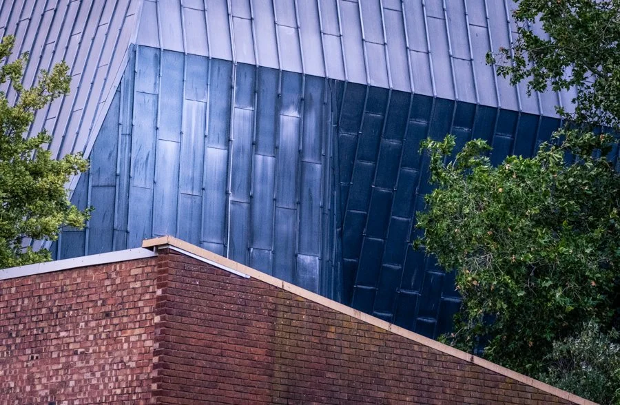 Angular brick and metal building surfaces meeting at contrasting angles, framed by surrounding trees in Coventry city centre.
