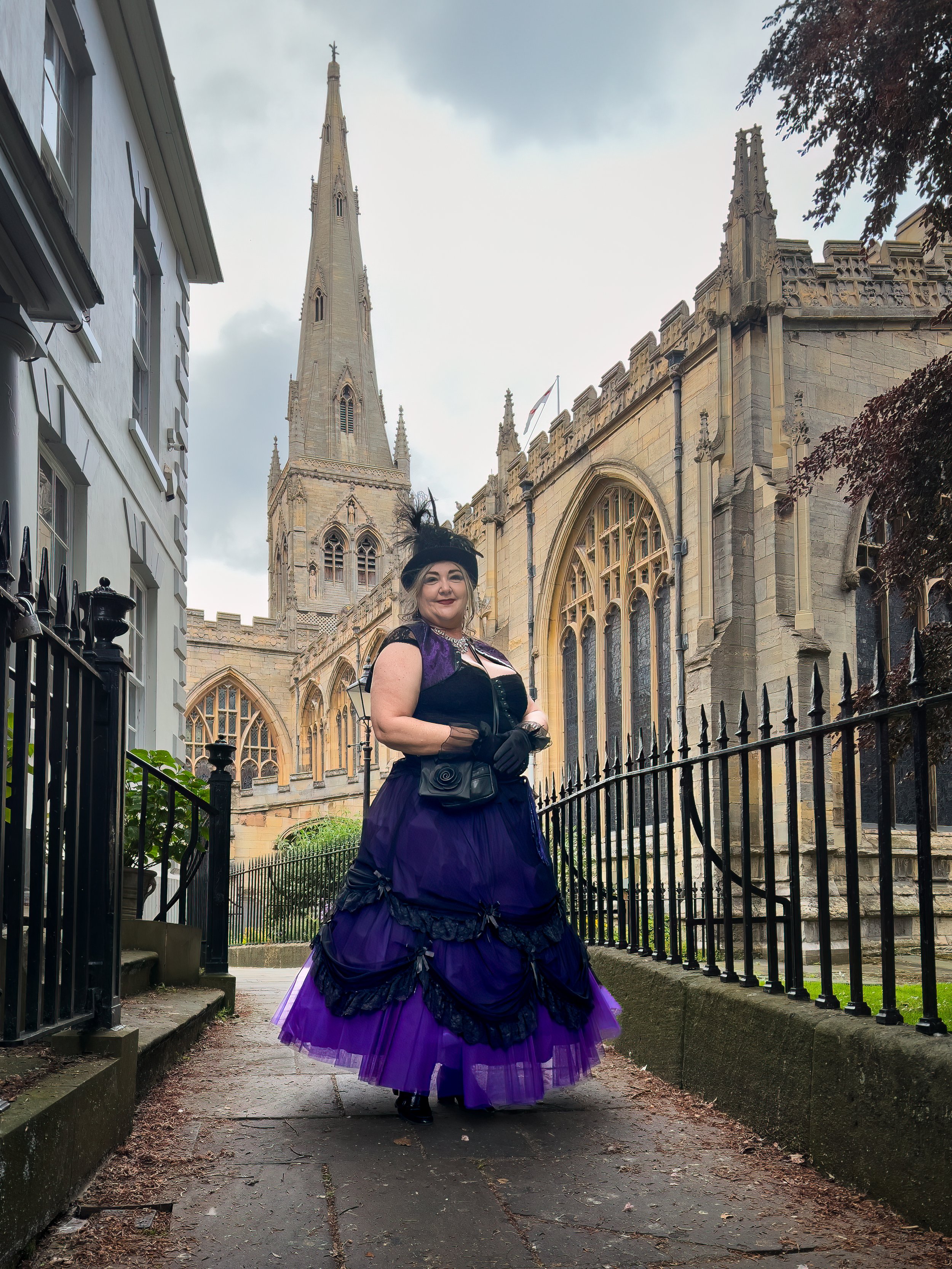 Portrait of Catherine Knee in a purple and black Victorian-style gown, posing beside a Gothic church with a tall stone spire. Dark clouds overhead and historic architecture create a dramatic, atmospheric setting.