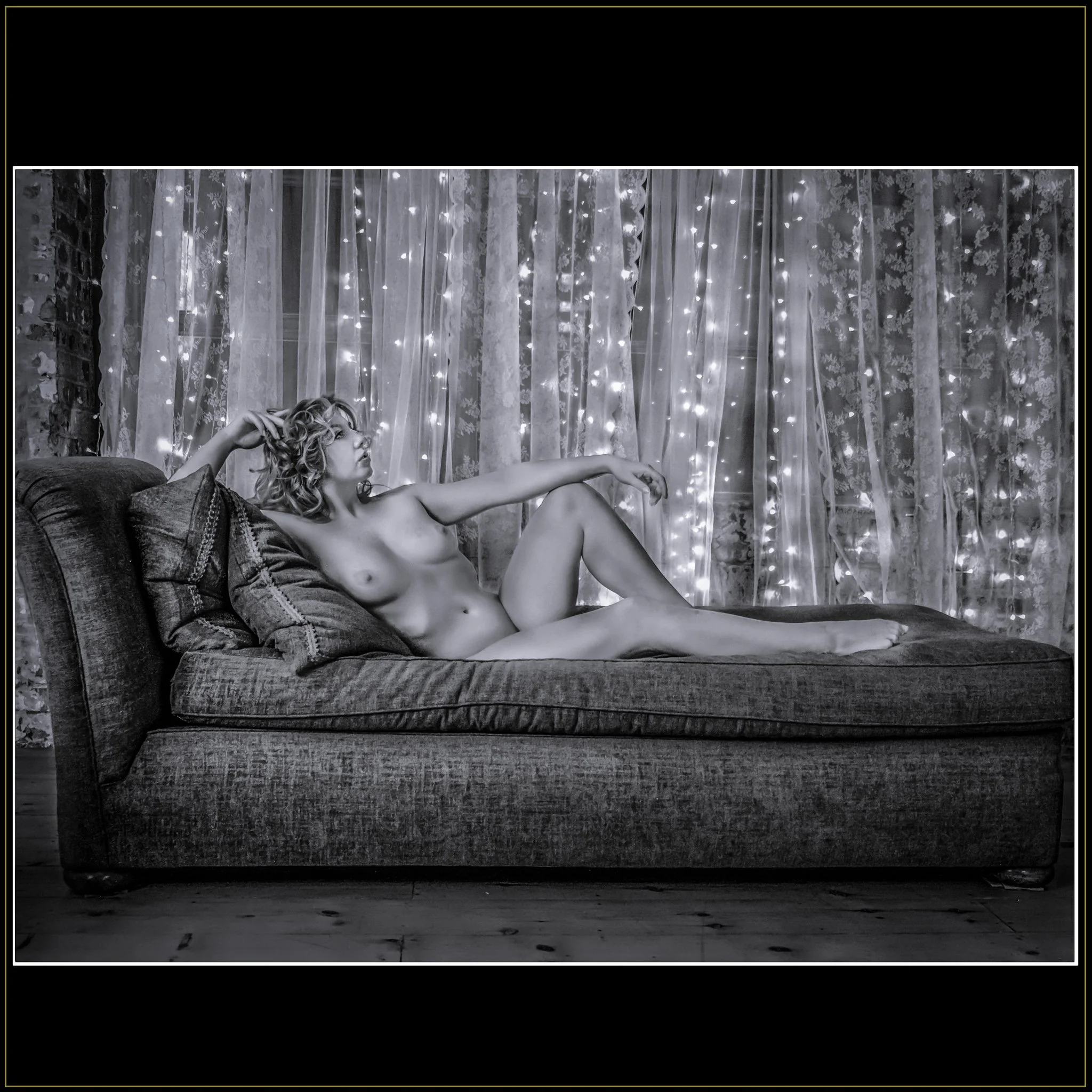 Reclining Nude – Fine Art Nude Photography by Catherine Knee. A nude female figure with curly hair lounges across a chaise longue, set against sheer curtains lit with strands of glowing fairy lights.
