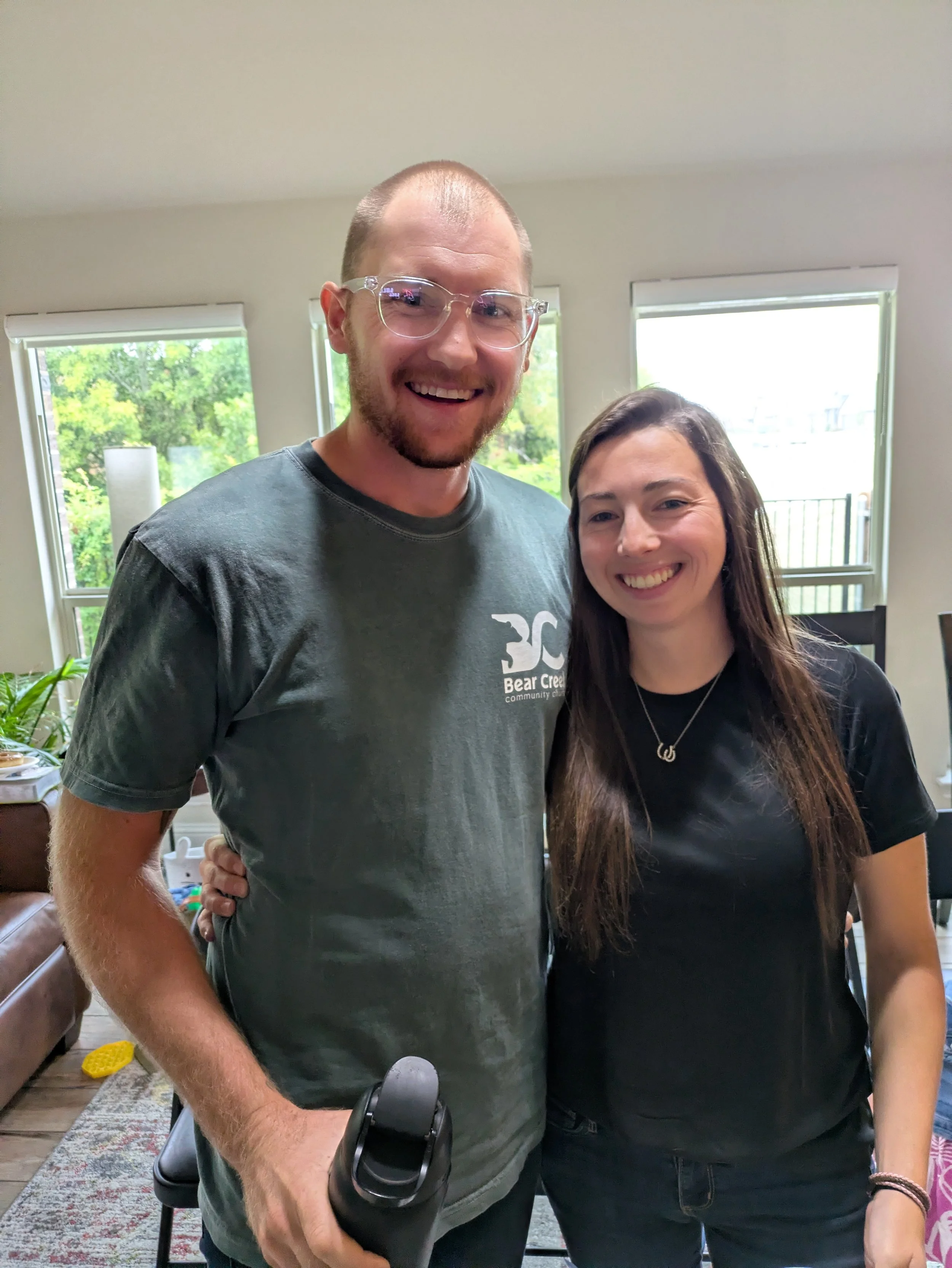 A man with glasses and a beard wearing a gray t-shirt with a graphic logo, smiling, standing next to a woman with long dark hair, smiling, in a bright room with windows showing greenery outside.