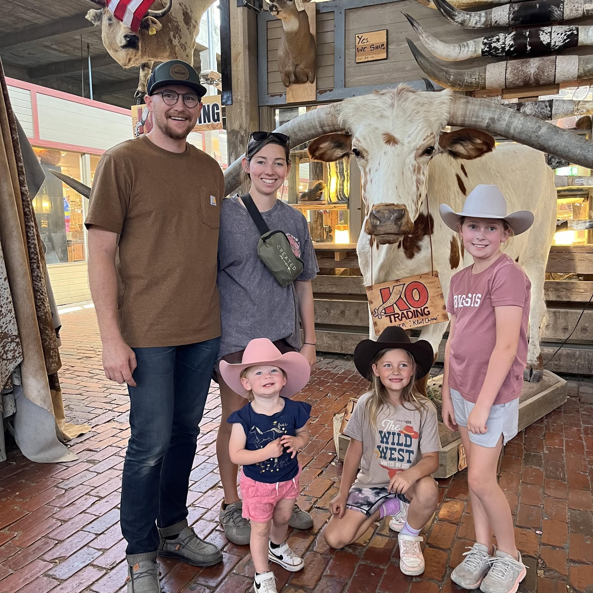 A group of five people, including two children, stands inside a store with mounted animal heads on the walls, including a large cow with long horns. They are smiling and posing for the photo. There are signs and various animal trophies in the backgro