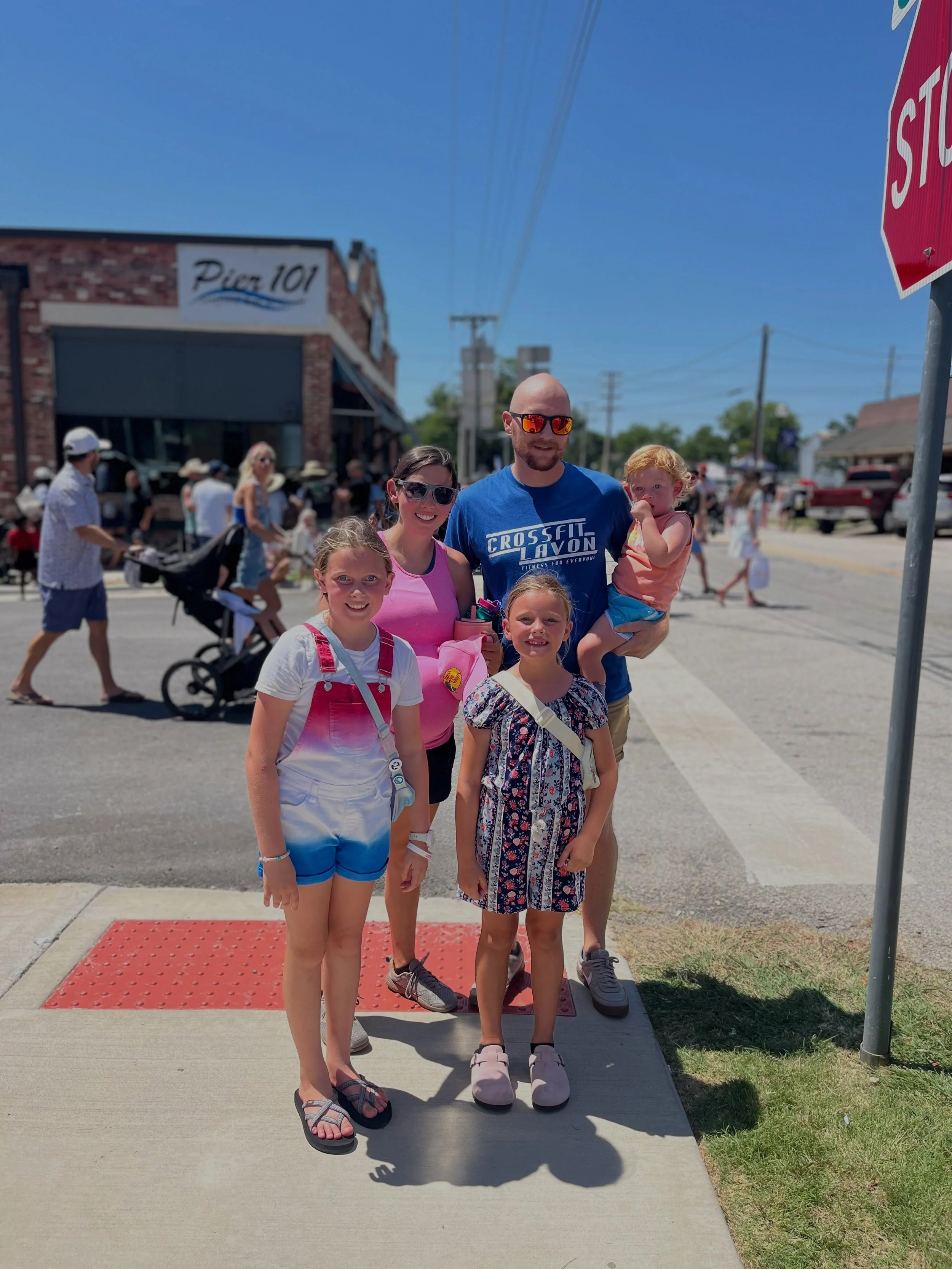 A group of five people, including three children and two adults, standing on a sidewalk during daytime. The children are in the front, one with blonde hair wearing a white shirt with pink overalls, and another with darker hair in a floral dress. The 