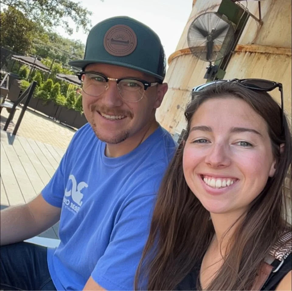 Smiling man and woman sitting outdoors on a wooden deck, with potted plants and a large beige structure in the background.