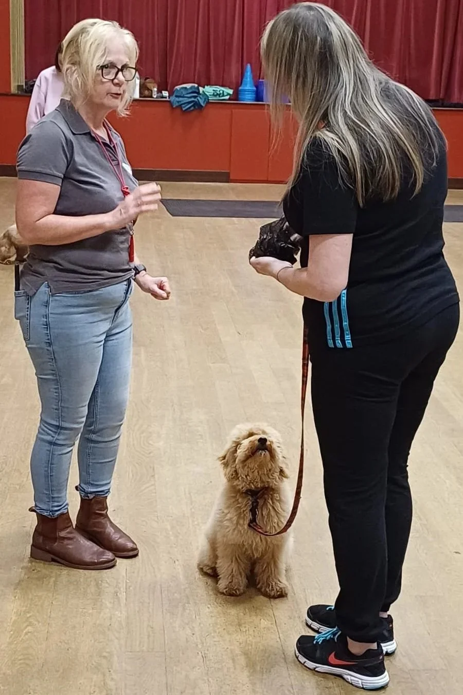 Two women and a small fluffy dog in an indoor training room. The woman on the left has blonde hair, glasses, and is wearing a gray polo shirt and jeans. The woman on the right has long blonde hair, is wearing a black shirt and black pants, and is holding the dog's leash. The dog sits attentively between them, looking up.