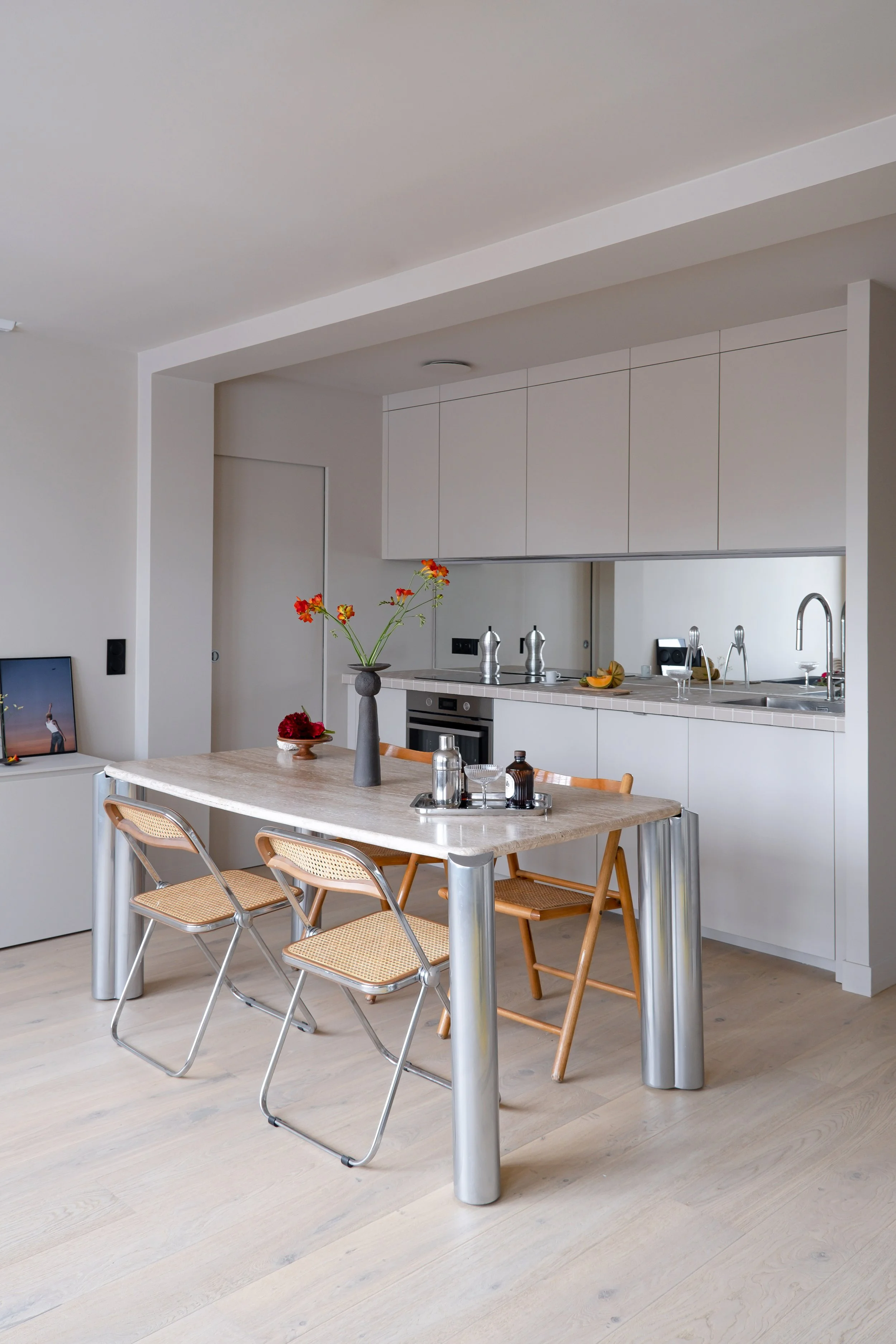 La table en marbre aux pieds cannelés en aluminium brossé occupe le centre de la pièce. Autour d'elle, des chaises pliantes en cannage et métal. La cuisine s'inscrit en fond, par un bandeau de placards en haut et en bas, sans poignées. Une crédence m