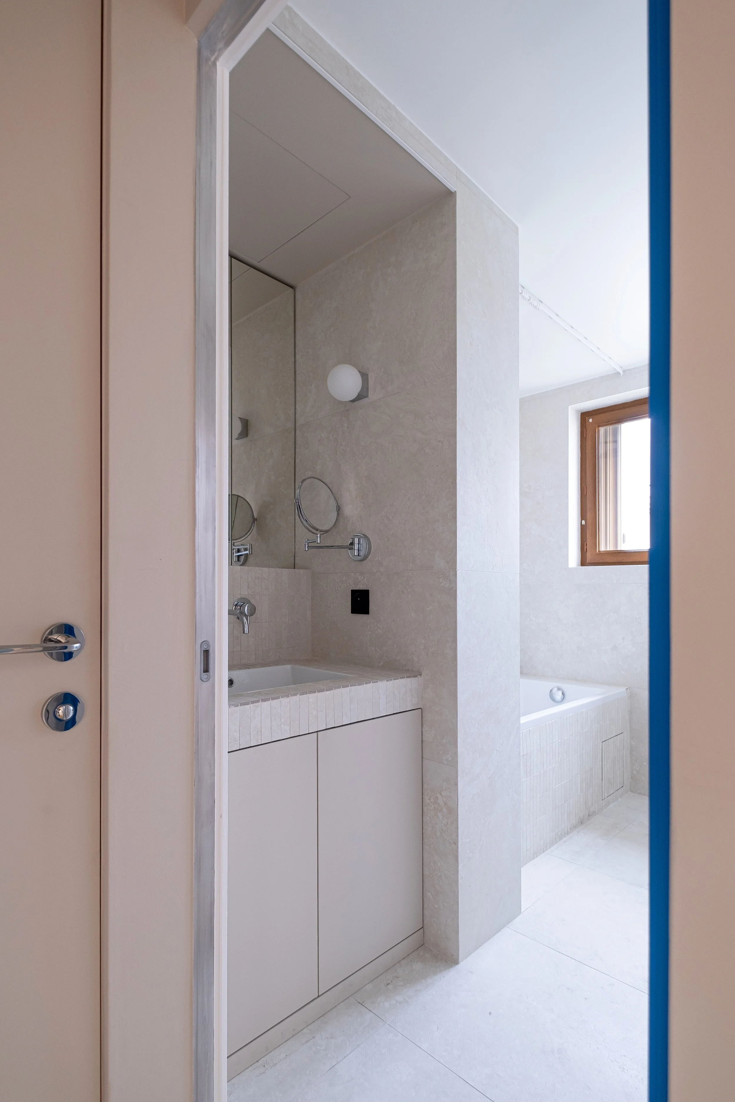 La salle de bain est vue depuis le couloir. La tranche bleue de la porte cadre l'entrée de la salle de bain. C'est un des traits architecturaux forts de cette pièce. À gauche, la vasque encastrée dans sa niche minérale. À droite, la baignoire est bai