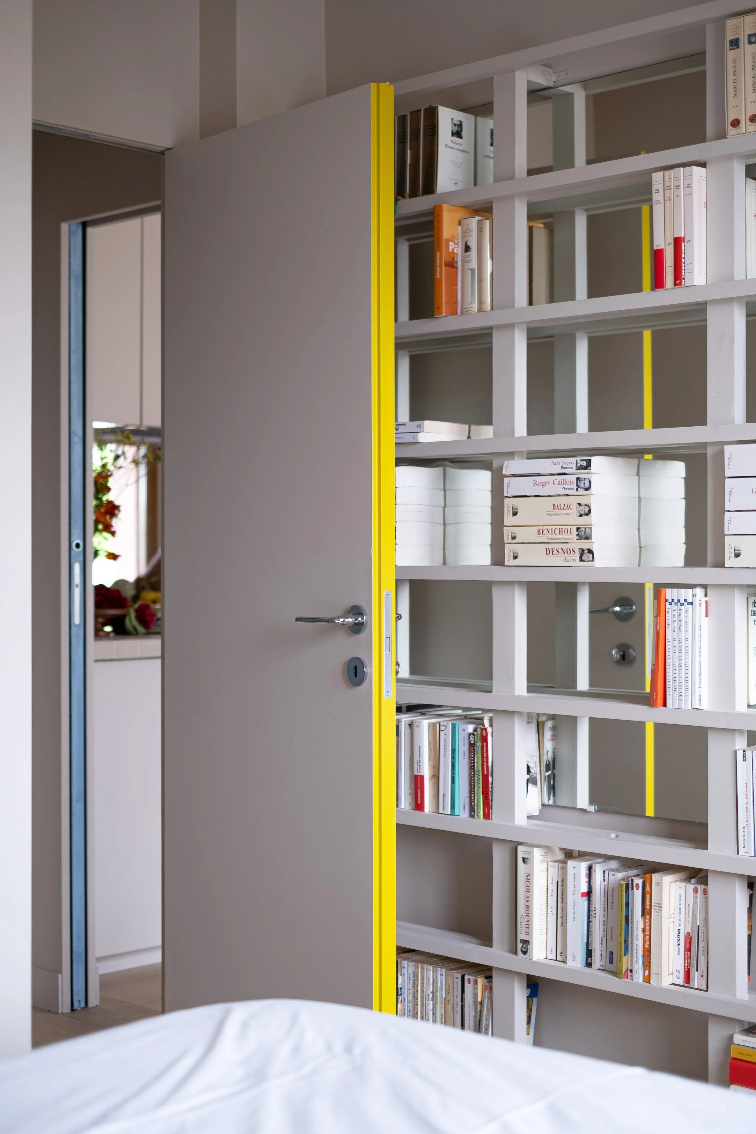 La tranche jaune de la porte révèle ici toute sa puissance. Fermée, elle disparaît dans le mur. Ouverte, elle devient un trait de couleur vif qui accentue l'environnement au coloris beige de la pièce. La bibliothèque design court du sol au plafond. L