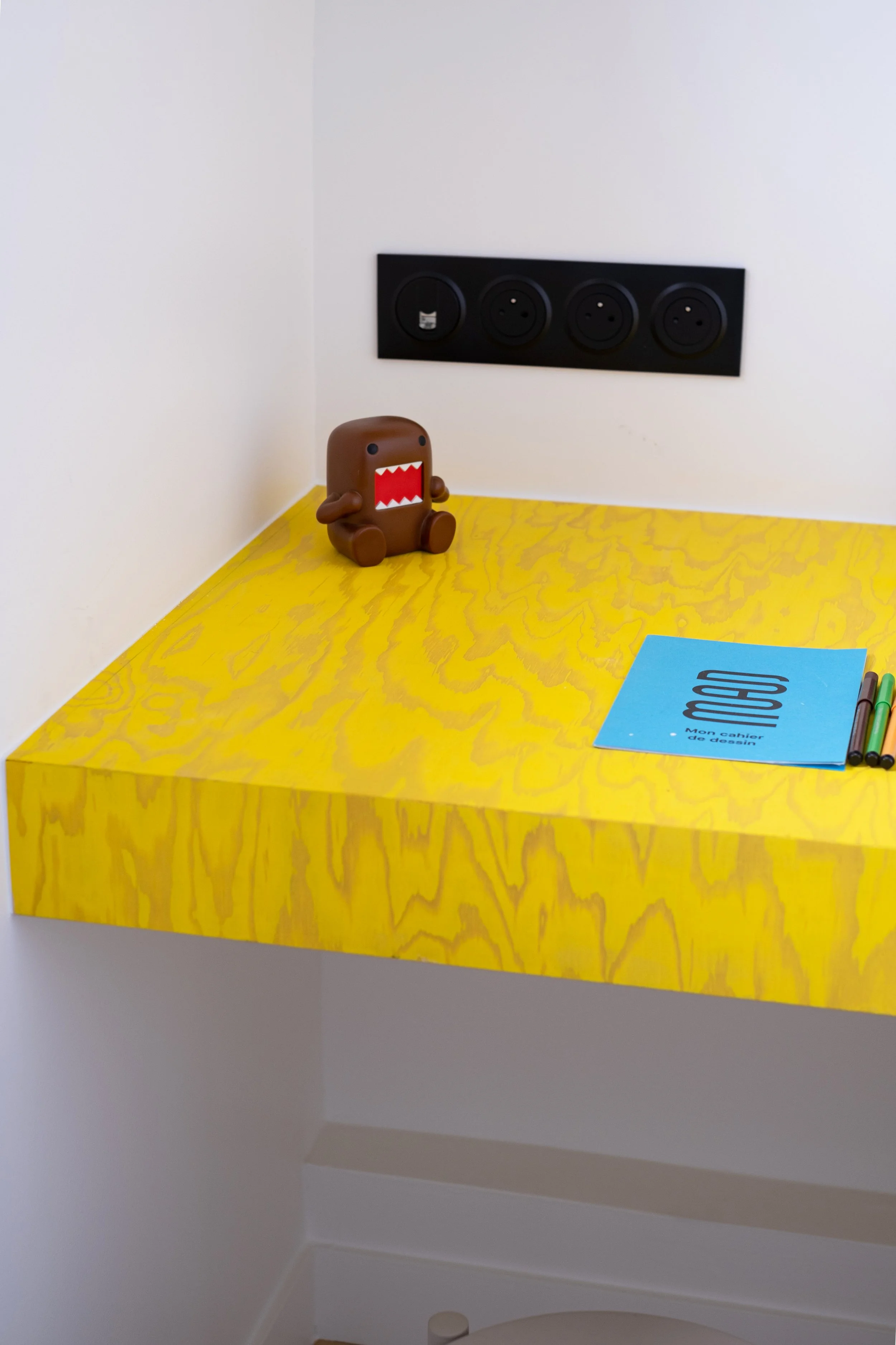 Voici un détails du bureau: la lasure jaune révèle le veinage du bois.  La texture est très graphique . Un cahier de dessin, quelques feutres, une figurine sont posés sur le bureau. Le bandeau de prises noir mat court en fond