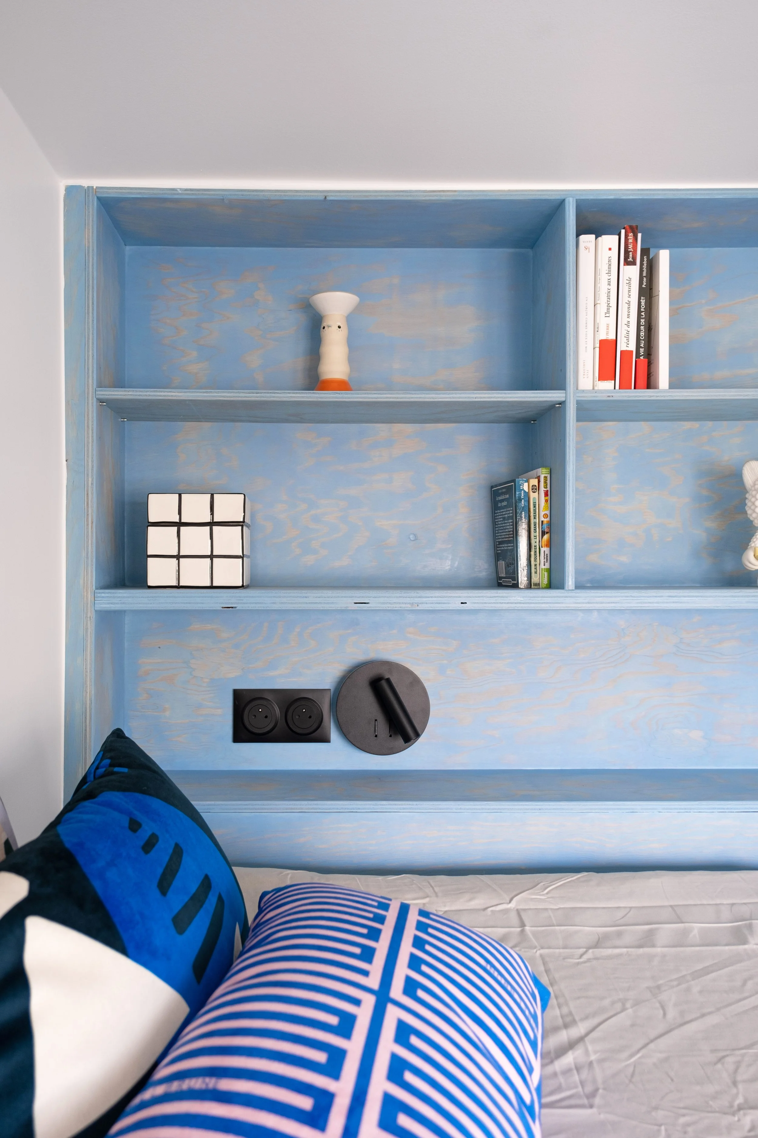 De près, la lasure bleue révèle le veinage du bois. C'est la même texture graphique que le bureau jaune, dans un coloris bleu. Sur les étagères, des objets de design, quelques livres, une petite figurine de designer. La prise noire et la liseuse rond