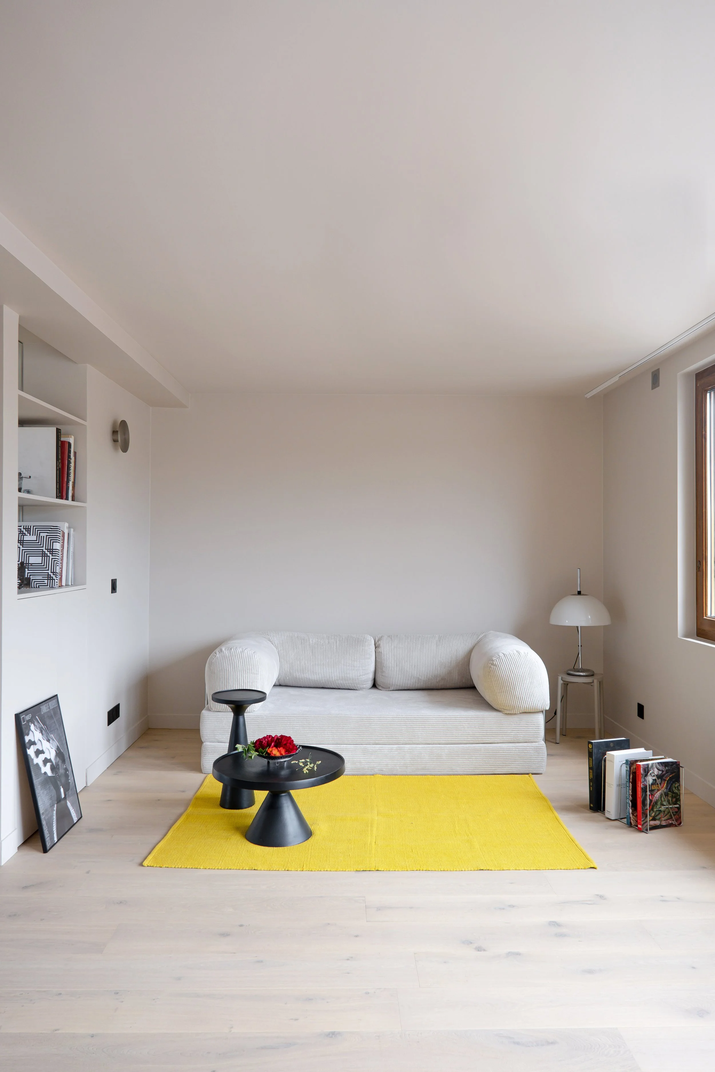Le canapé en velours côtelé crème occupe le centre de la pièce. Sur le côté, on observe deux tables basses noires. Le tapis jaune vif tranche dans l'ensemble beige et blanc, une belle touche de couleur assumée. À gauche, une niche intégrée accueille 
