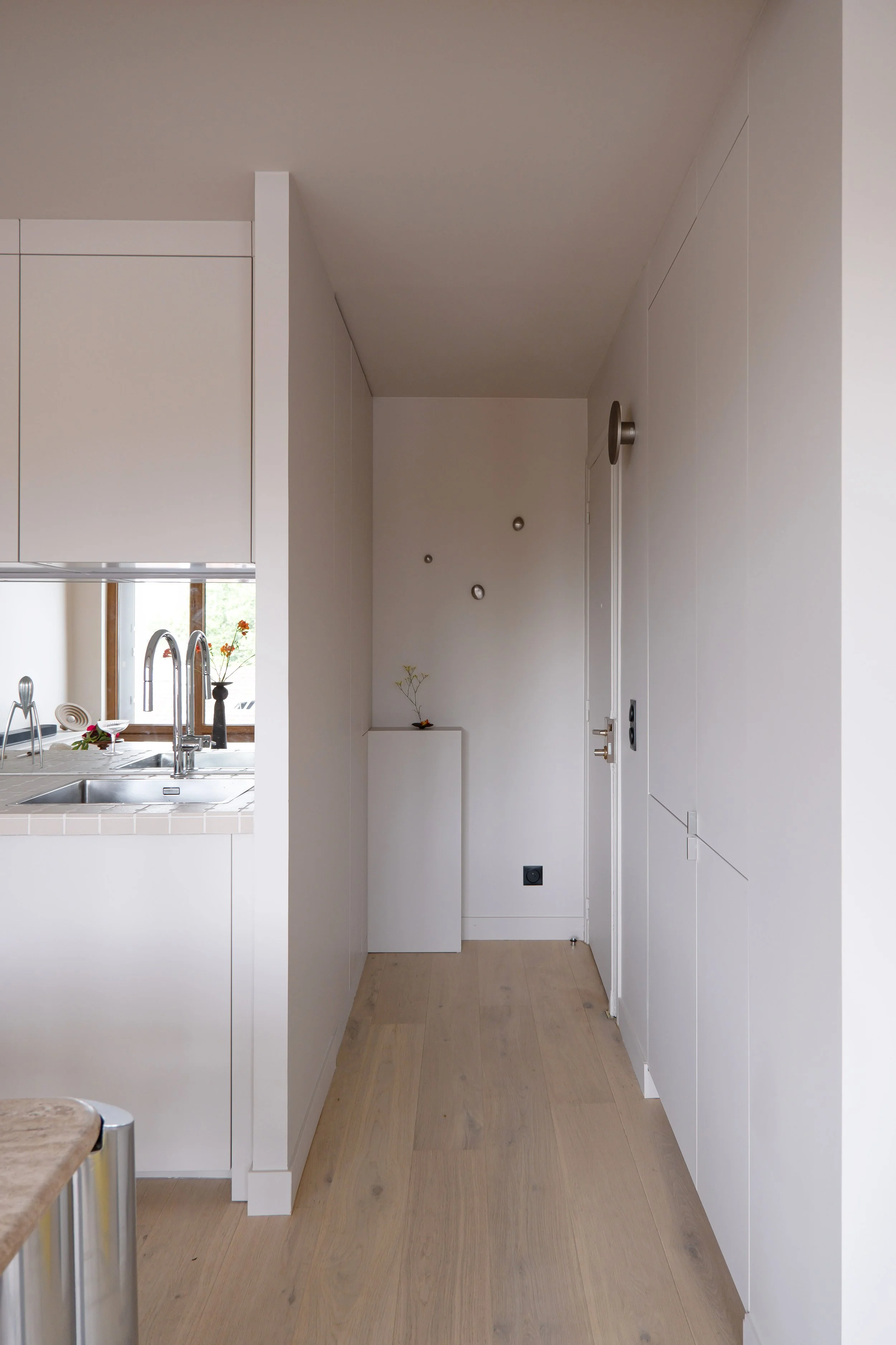 L'entrée s'ouvre sur un couloir épuré, structuré par des rangements intégrés, tous à fleur de mur. Au fond, une sellette blanche accueille un vase et une branche et pour y poser ses clefs, un endroit minimaliste et fonctionnel qui marque le seuil. La