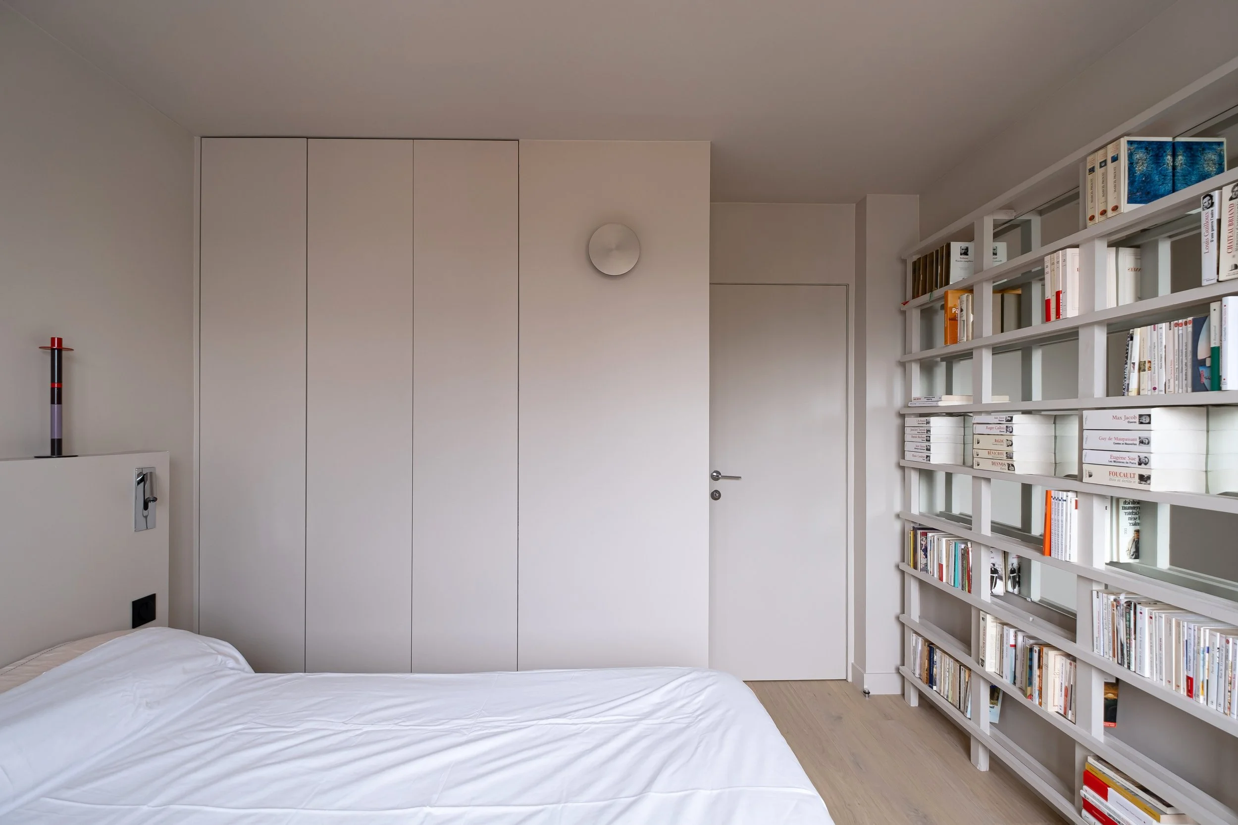 Face au lit, un mur de placards sans poignées est placé sur toute la hauteur du mur. Les détails sont minimalistes pour un cachet discret et architectural. La porte s'y fond naturellement. Sur la droite, la bibliothèque sur mesure habille tout le mur