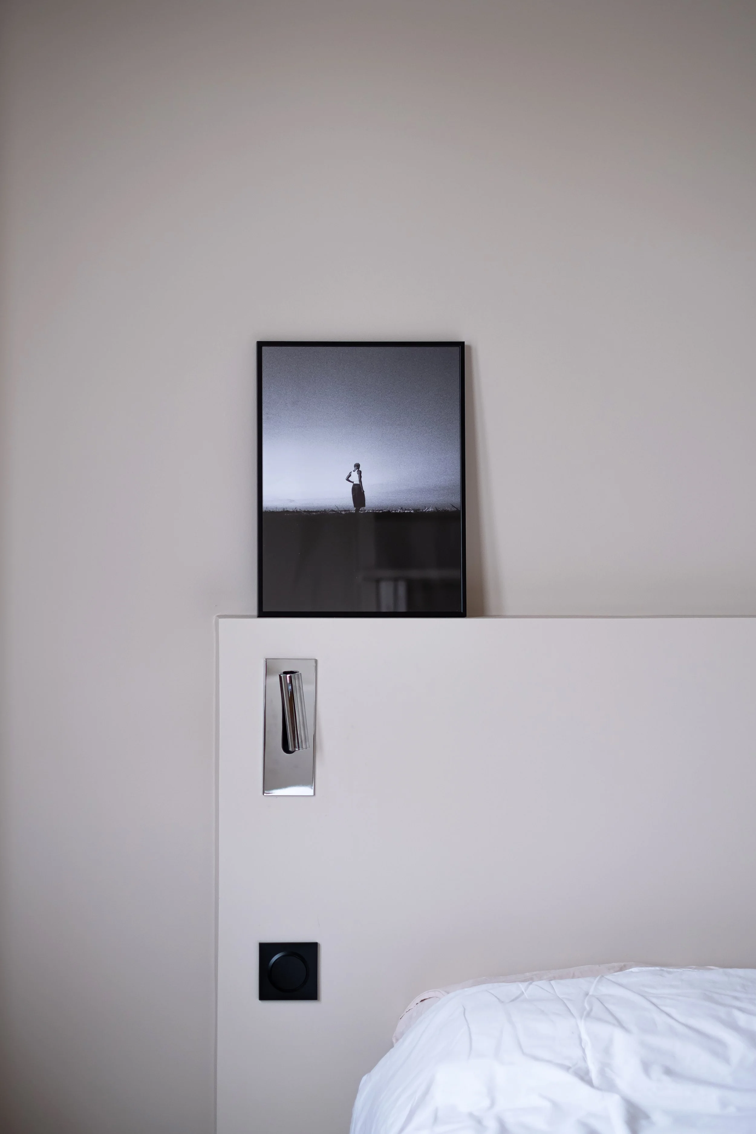 La liseuse chromée s'encastre dans la tête de lit sur mesure. Sur le rebord, une photographie en noir et blanc est simplement posée.  En dessous, il y a un interrupteur noir mat et intégré à la tête de lit. Cette composition est précise et discrète.