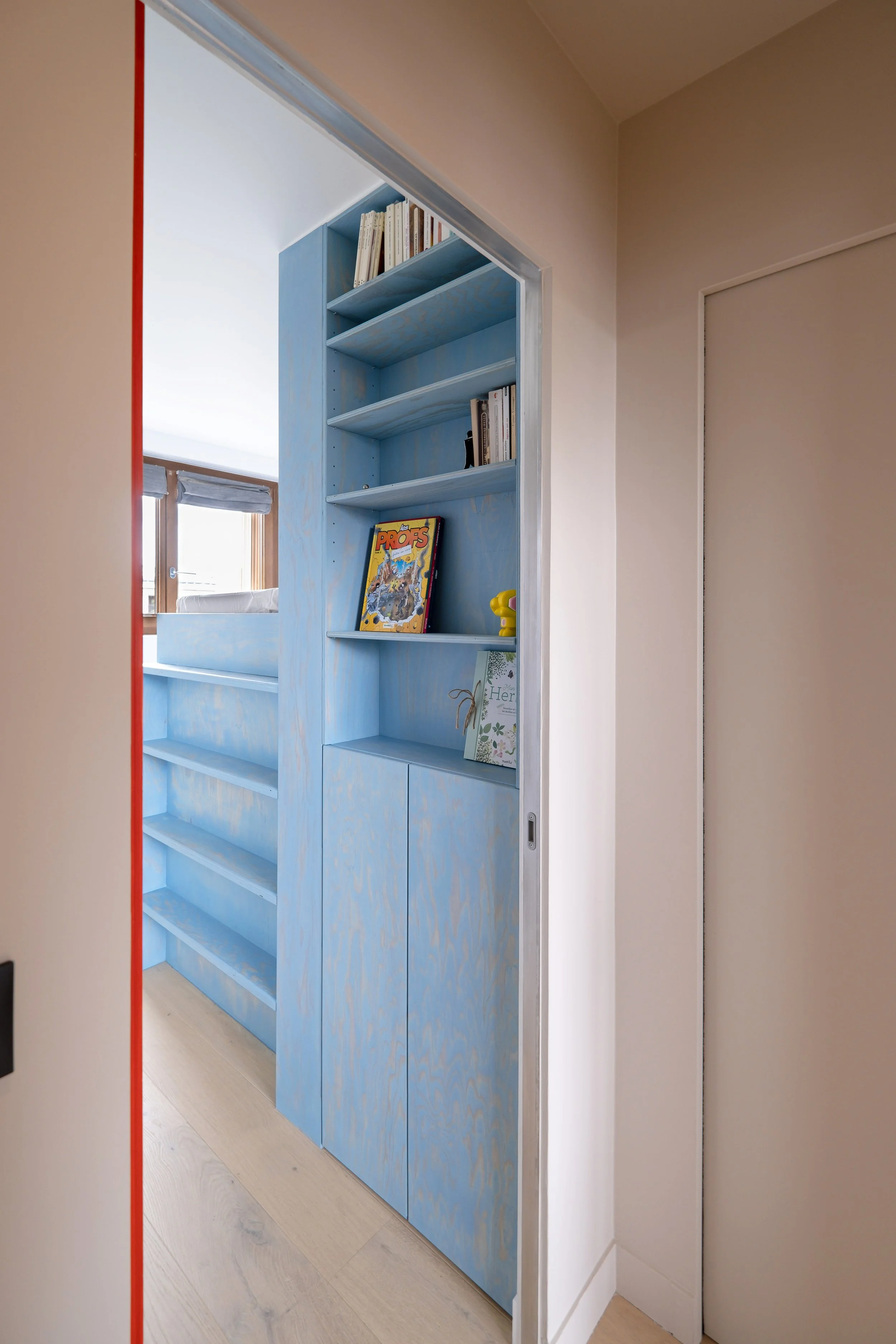 Depuis le couloir, la chambre se révèle par fragments. La bibliothèque bleue s'inscrit dans l'encadrement de la porte comme un tableau. En bas, des placards fermés. En haut, des étagères ouvertes avec des livres et des objets de décoration. On peut v