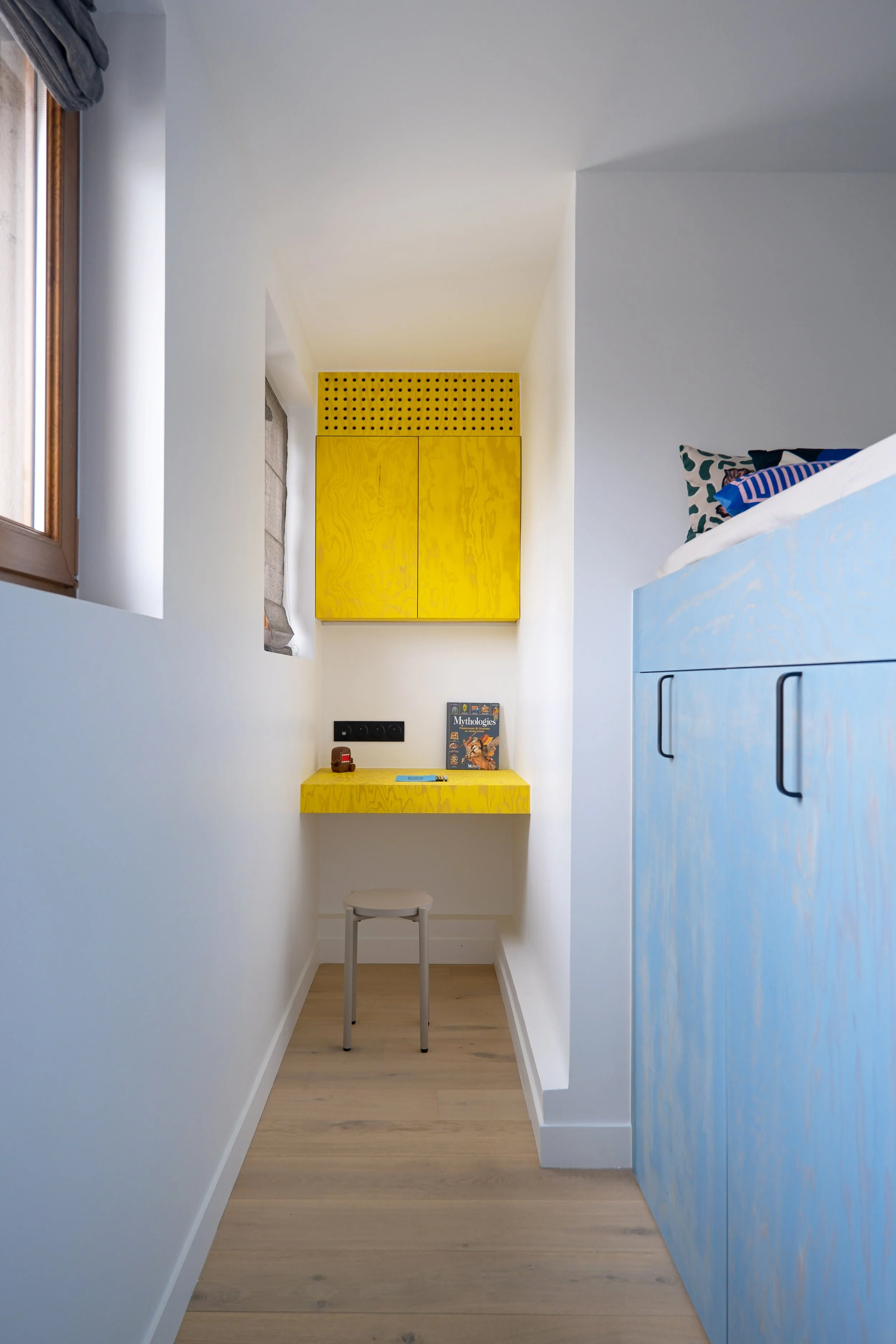 Dans une alcôve qui se situe entre le lit surélevé et la fenêtre, se trouve un bureau en bois lasuré, coloris jaune. Au-dessus du plan de travail, un placard aux portes perforées. Un tabouret métallique est glissé sous le bureau et un livre est posé 