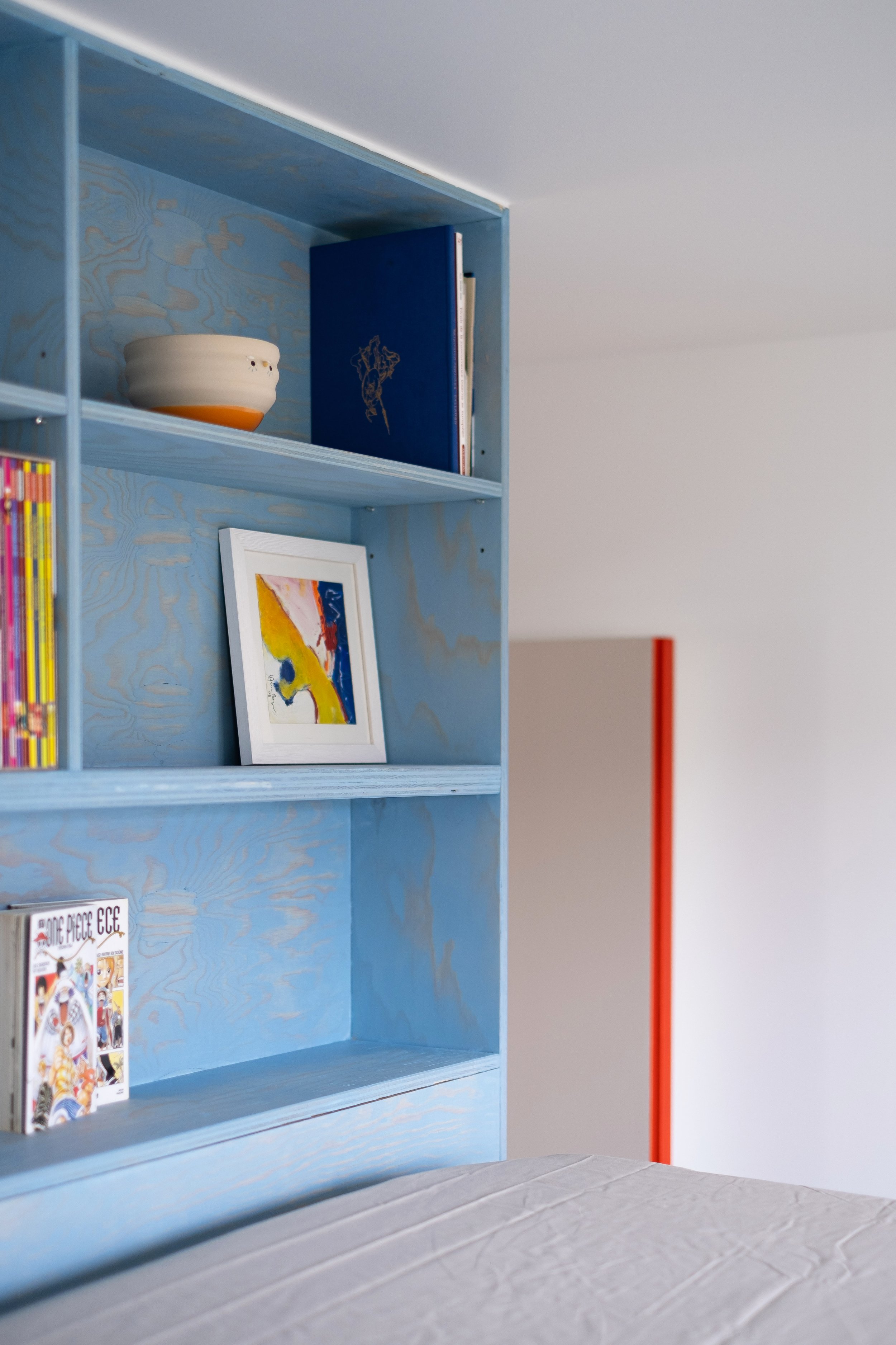 Sur les étagères en bois lasuré bleu se trouve une aquarelle, un bol en céramique, des mangas, un carnet bleu marine. En arrière-plan, la tranche rouge de la porte voisine apparaît sur la droite. Un trait de couleur qui rappelle que l'appartement ent