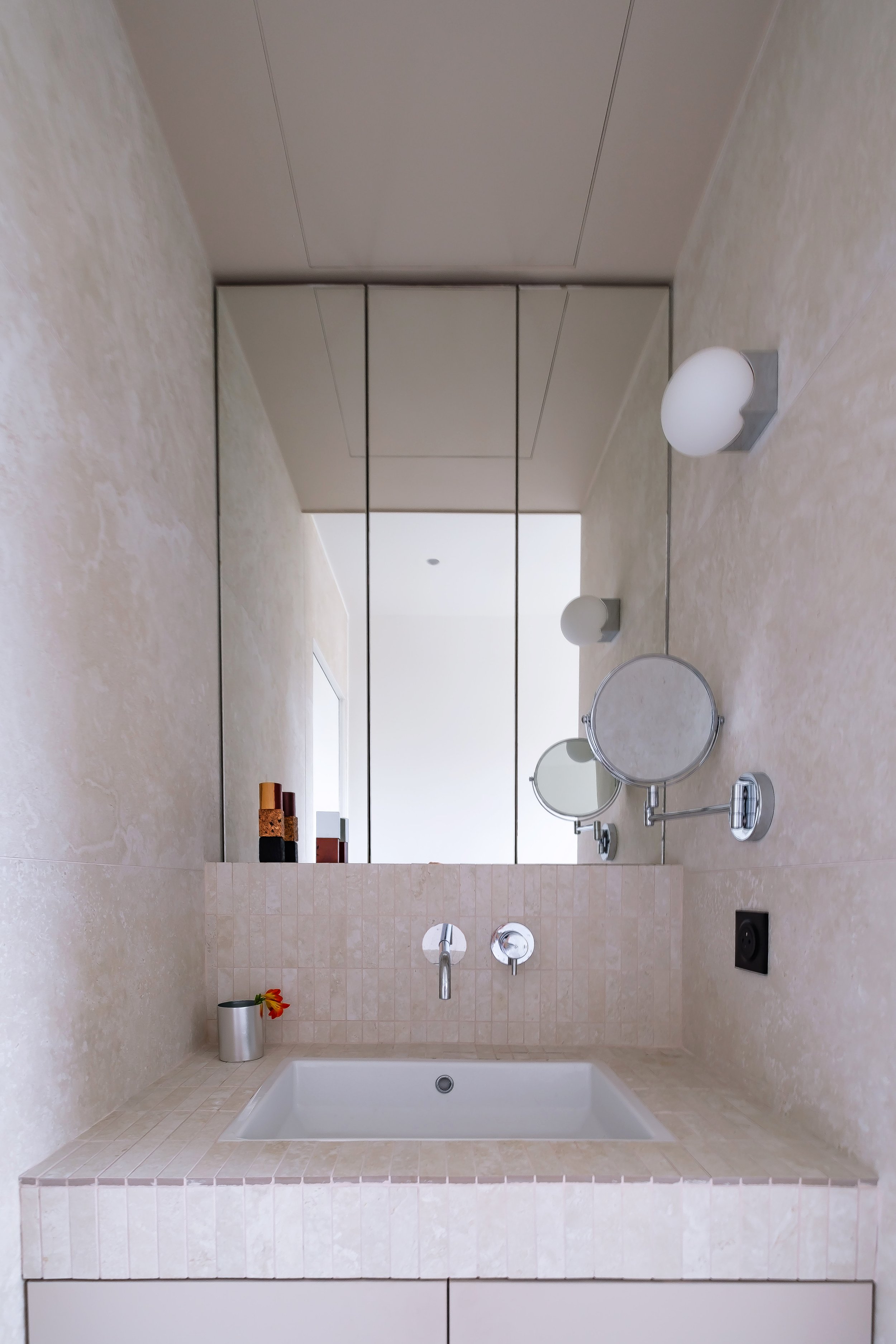 La vasque est encastrée dans un plan de travail entièrement habillé de petits carreaux en matière minérale. La robinetterie est encastrée dans le mur, chromée et minimaliste. Le miroir en triptyque occupe toute la largeur de la niche et remonte jusqu