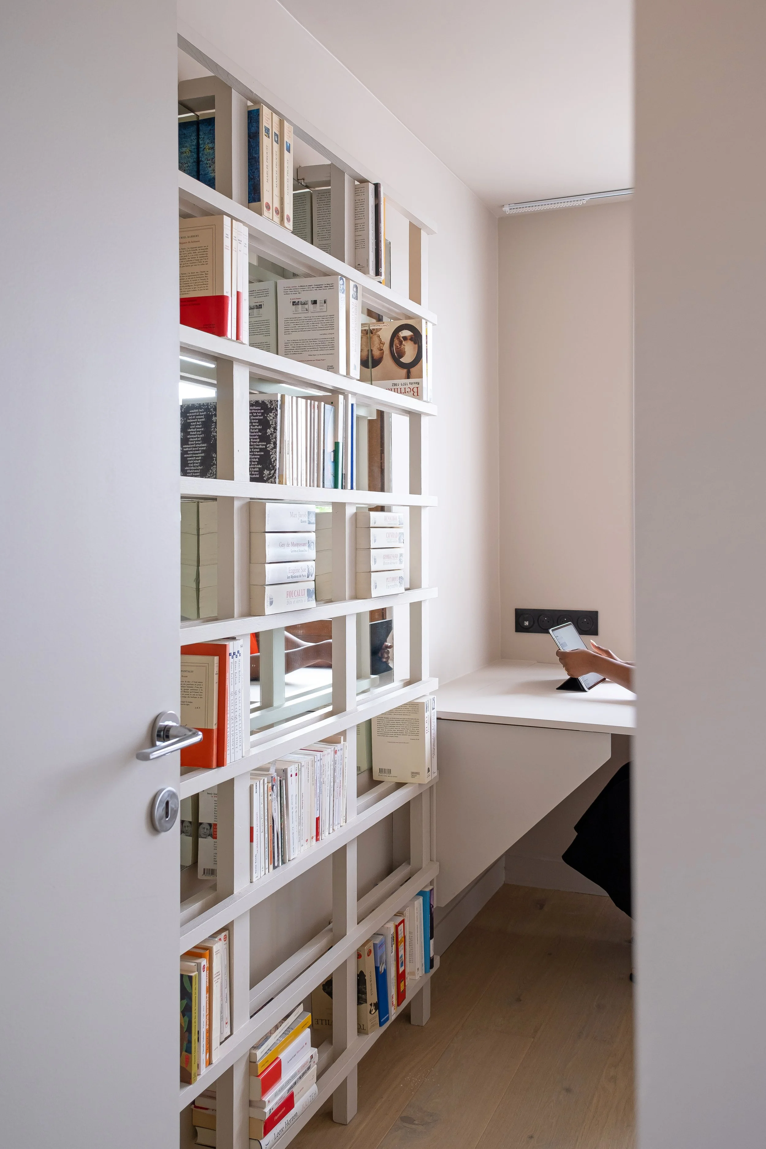 Derrière la bibliothèque, un bureau escamotable est accolé au mur. L'espace est ergonomique,  intime, et structuré pour être le plus efficace. Une main tient une tablette. La pièce, en plus d'être une chambre et une bibliothèque, trouve ici une trois
