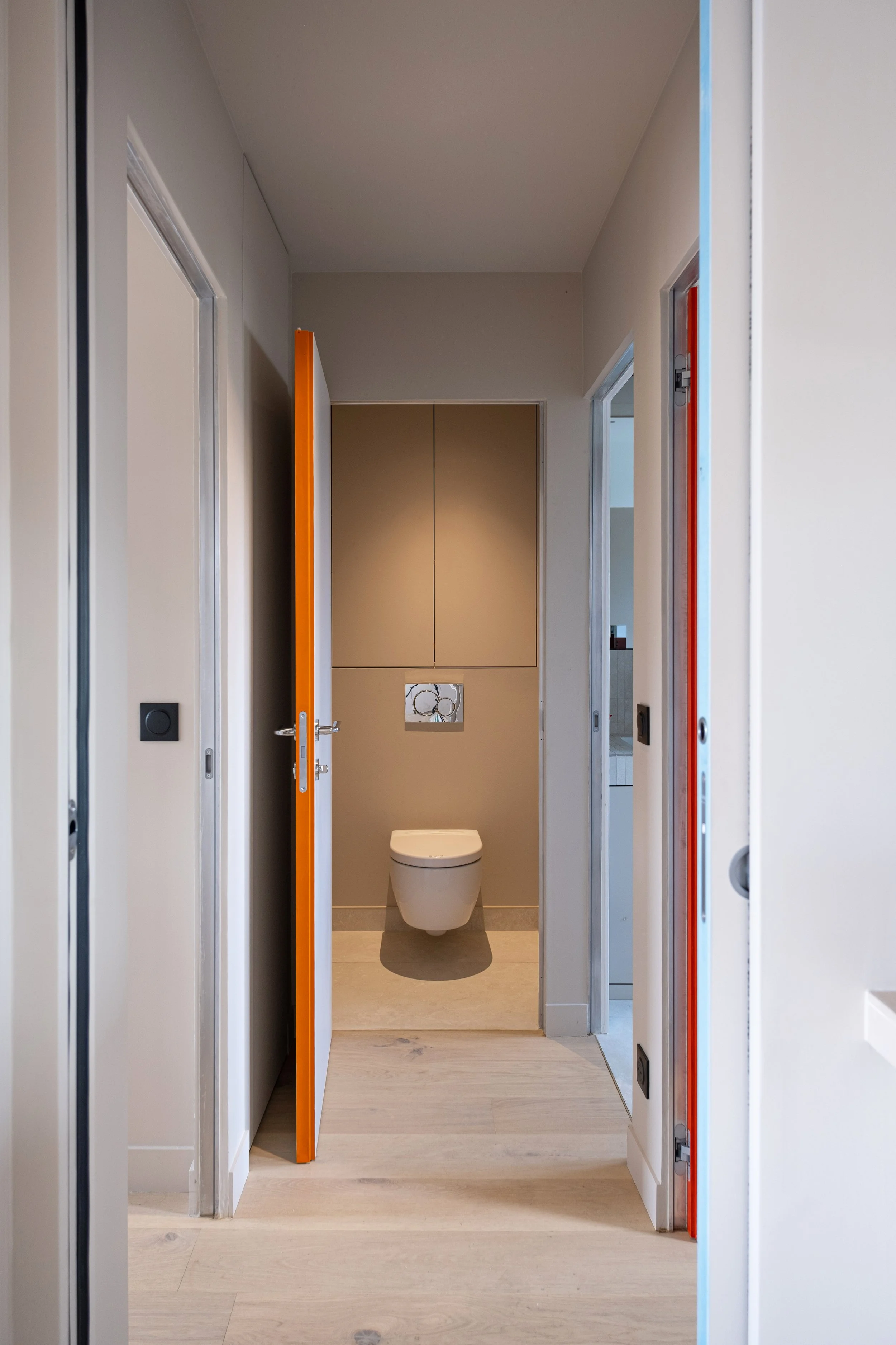 Le couloir distribue les pièces avec poésie. Au fond, on peut voir les WC suspendus, avec un placard sur mesure en teinte taupe au-dessus. La teinte générale est beige. Sur la gauche, on distingue la tranche orange d'une porte. Sur la droite, la tran