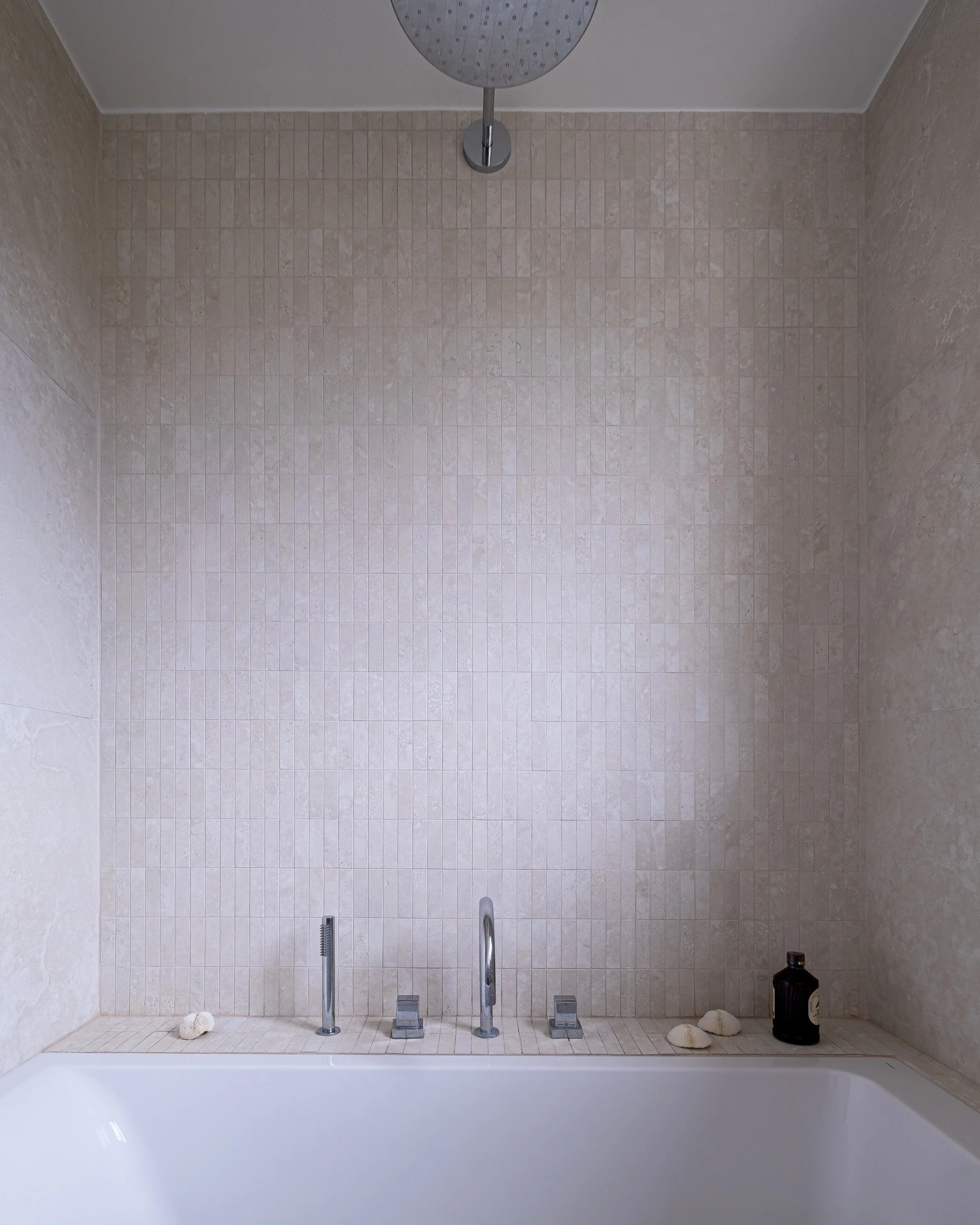 Le mur de petits carreaux, en texture minérale, monte du bord de la baignoire jusqu'au plafond, sur toute la surface du mur. Le ciel de pluie, rond et chromé, est centré au-dessus de la baignoire. En bas, la robinetterie de baignoire et quelques gale