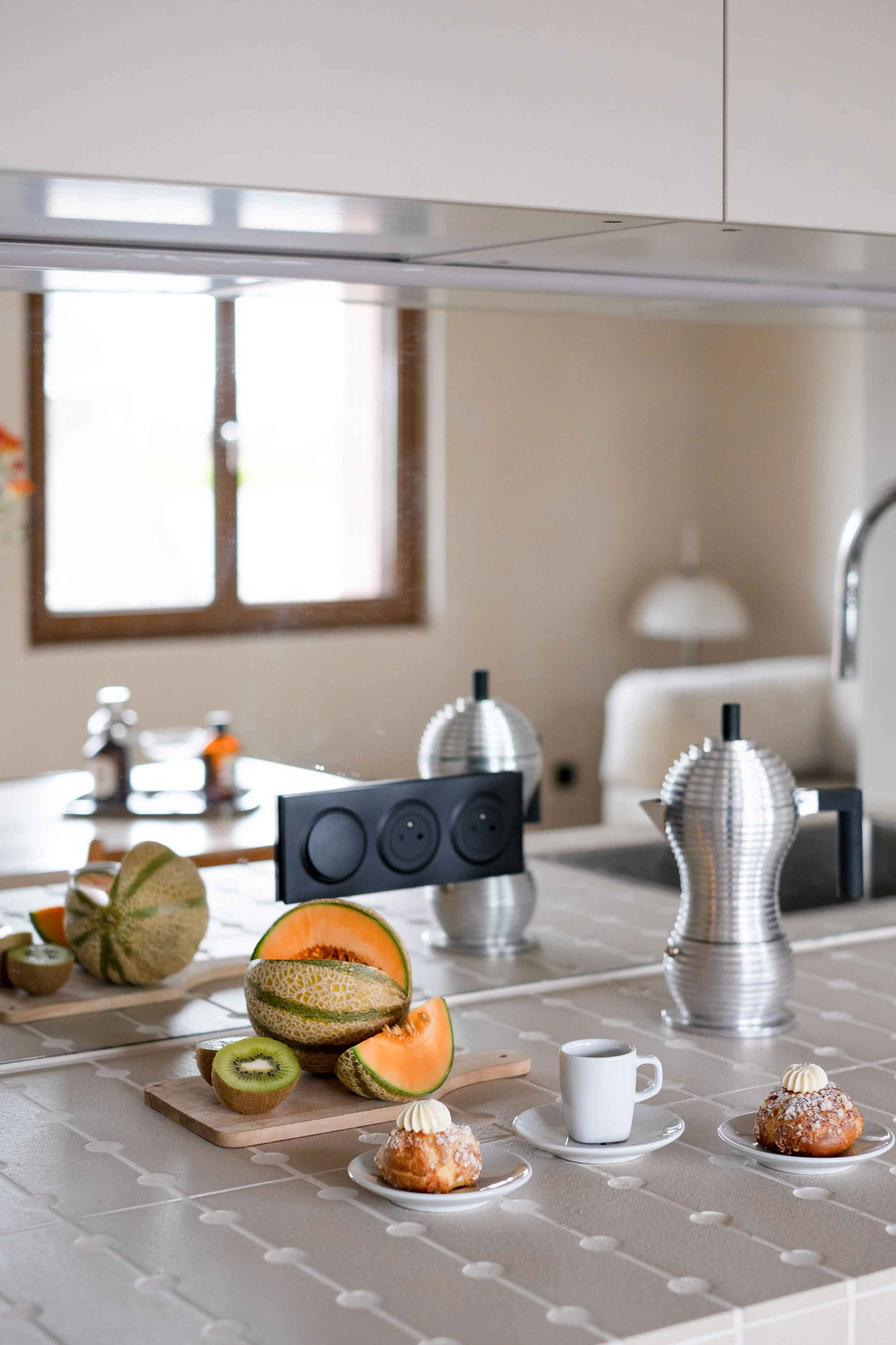 La cafetière Pulcina d'Alessi, pièce iconique dont les reflets inox dialoguent avec la crédence miroir, est posé sur le plan de travail en carreaux de grès beige. La prise noire mate, encastrée en bandeau, amène un accent à l'ensemble. Derrière, le m