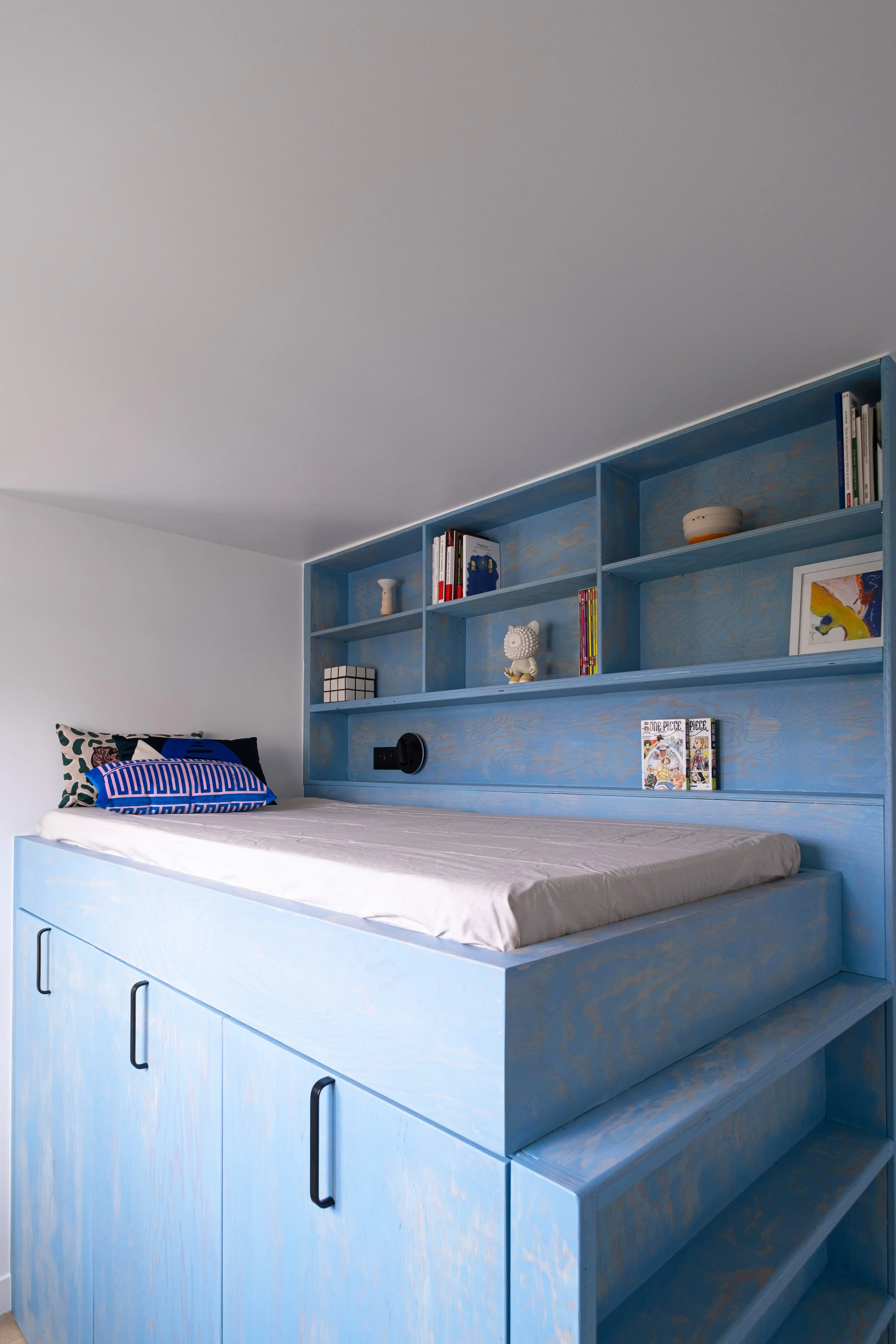 Le lit surélevé en bois lasuré bleu est une pièce de design à lui seul. En dessous, des placards coulissants à poignées noires dissimulent rangements et buanderie. Au niveau de la tête de lit, une bibliothèque intégrée court sur toute la longueur du 