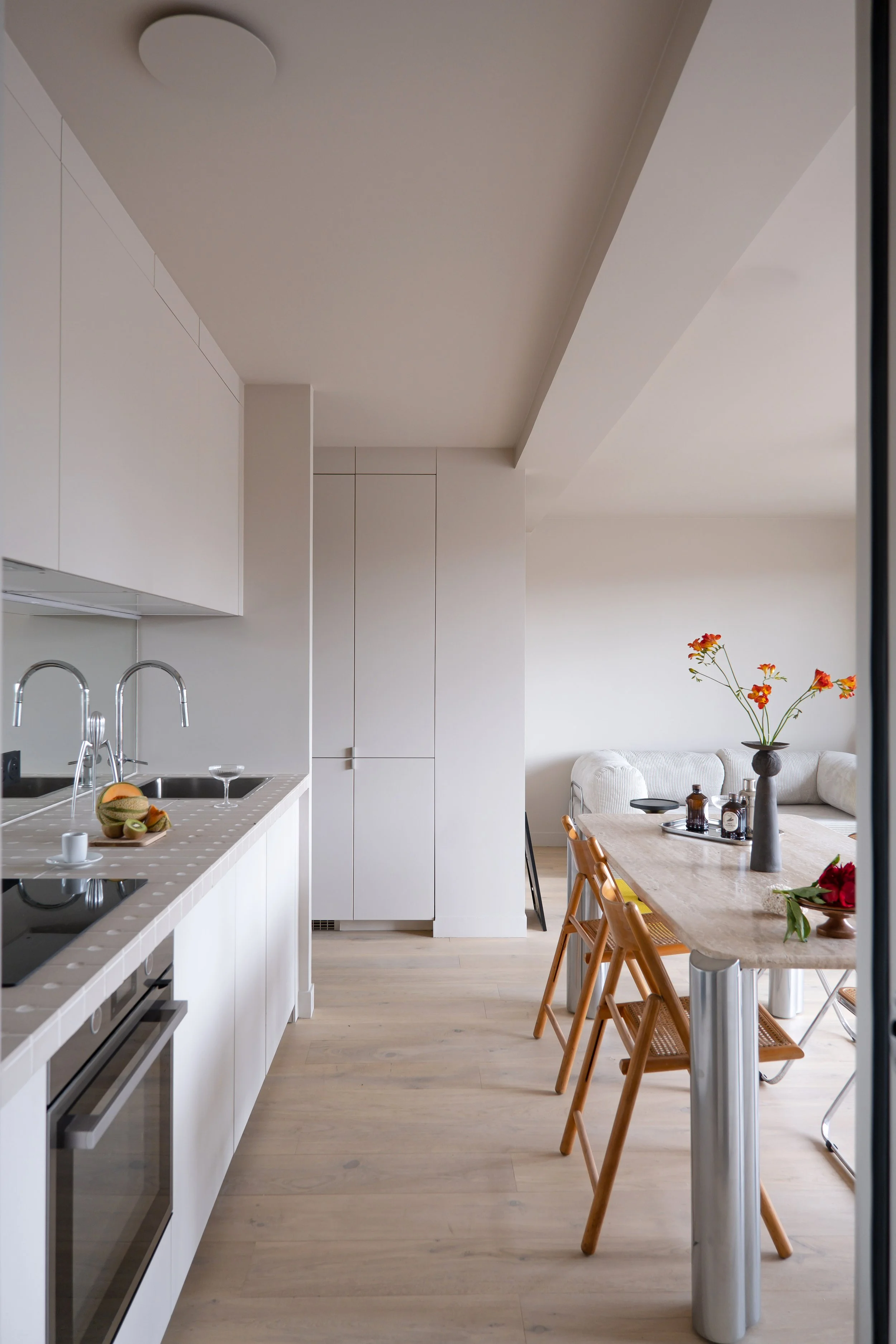 Dès le seuil, la cuisine se déploie toute en longueur. Les placards sans poignées s'alignent sur 2 rangées au niveau du sol et du plafond, unifiés dans un même beige mat. Le plan de travail en carreaux de grès court sur toute la longueur. Au fond, la