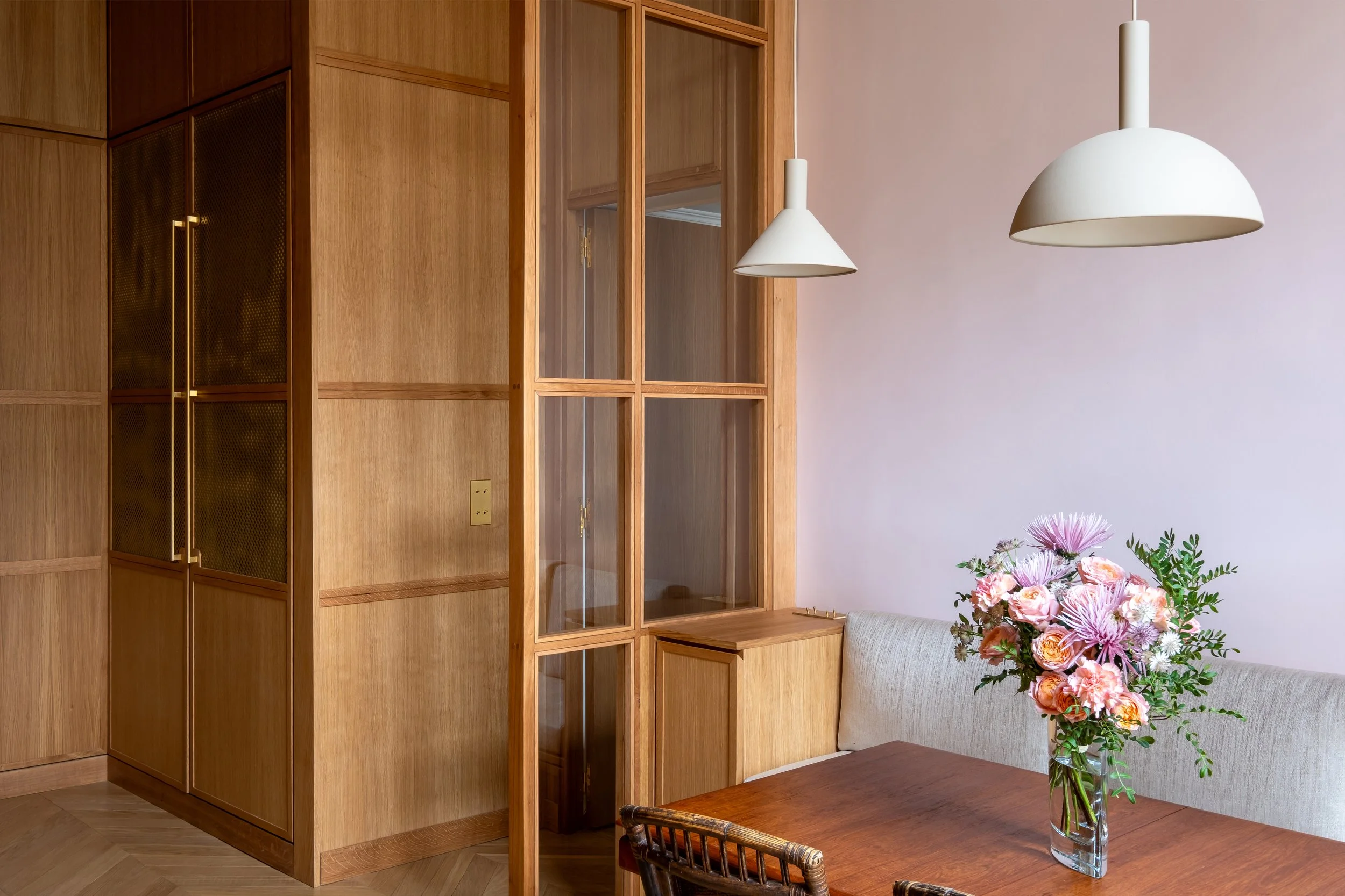 Verrière chêne sur mesure, coin repas séparé du garde-manger, suspensions asymétriques, poignées laiton tout en longueur, mur rose poudré, Paris.