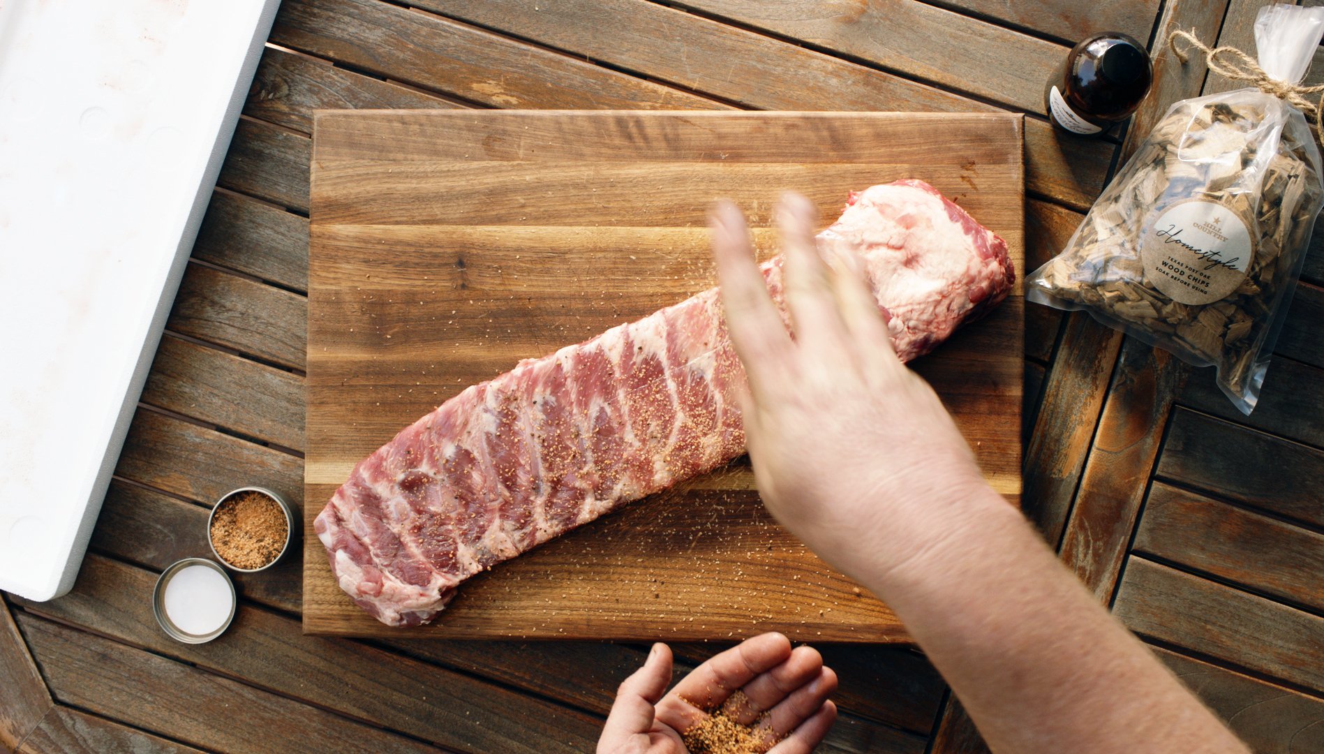 Person seasoning a rack of pork ribs on a wooden cutting board, with wood chips and a seasoning container nearby, on a wooden table.