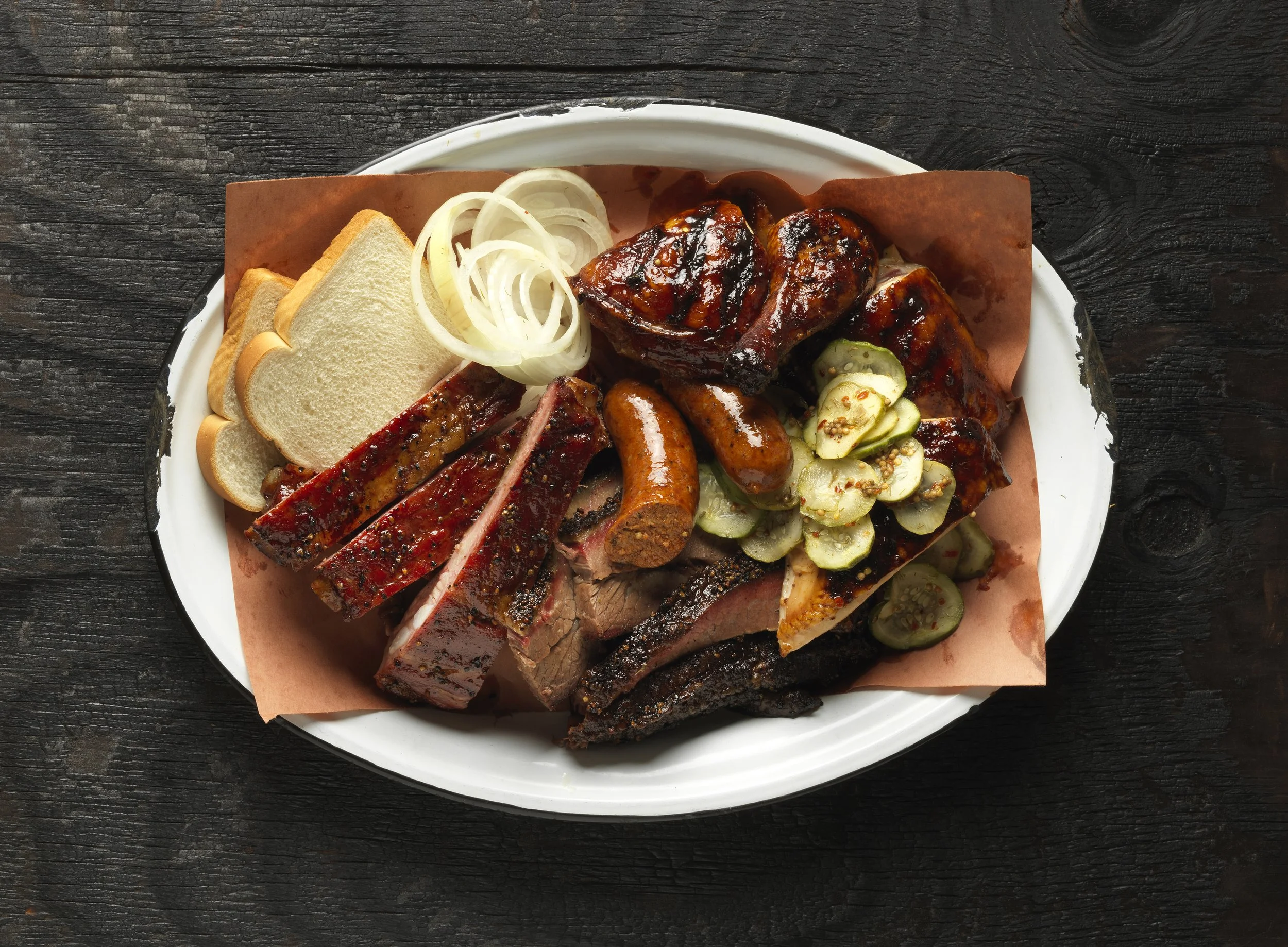 Platter of smoked barbecue meats with sliced bread, pickles, and onions on a white plate.