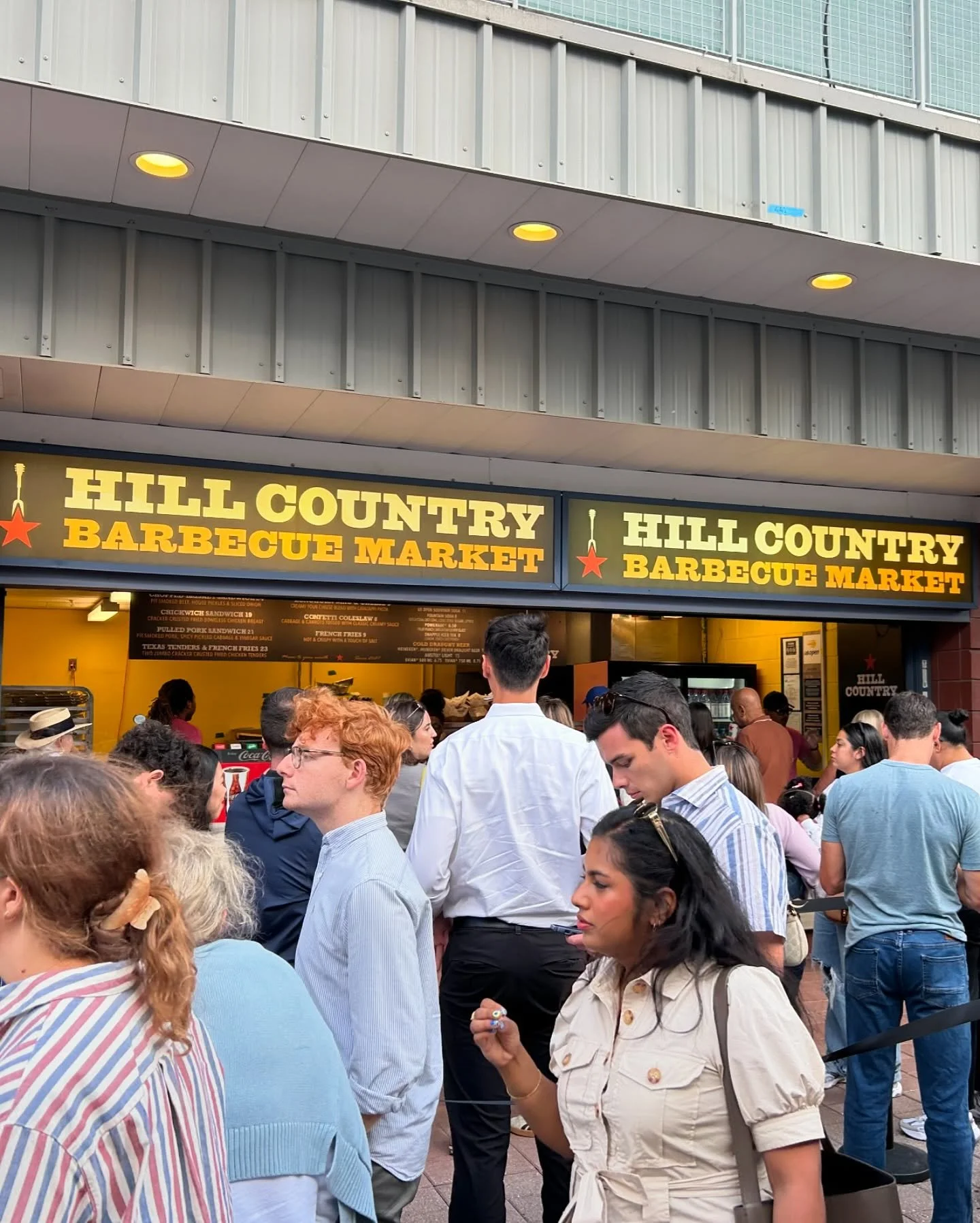 Crowd waiting outside Hill Country Barbecue Market with signage and visible menu options.