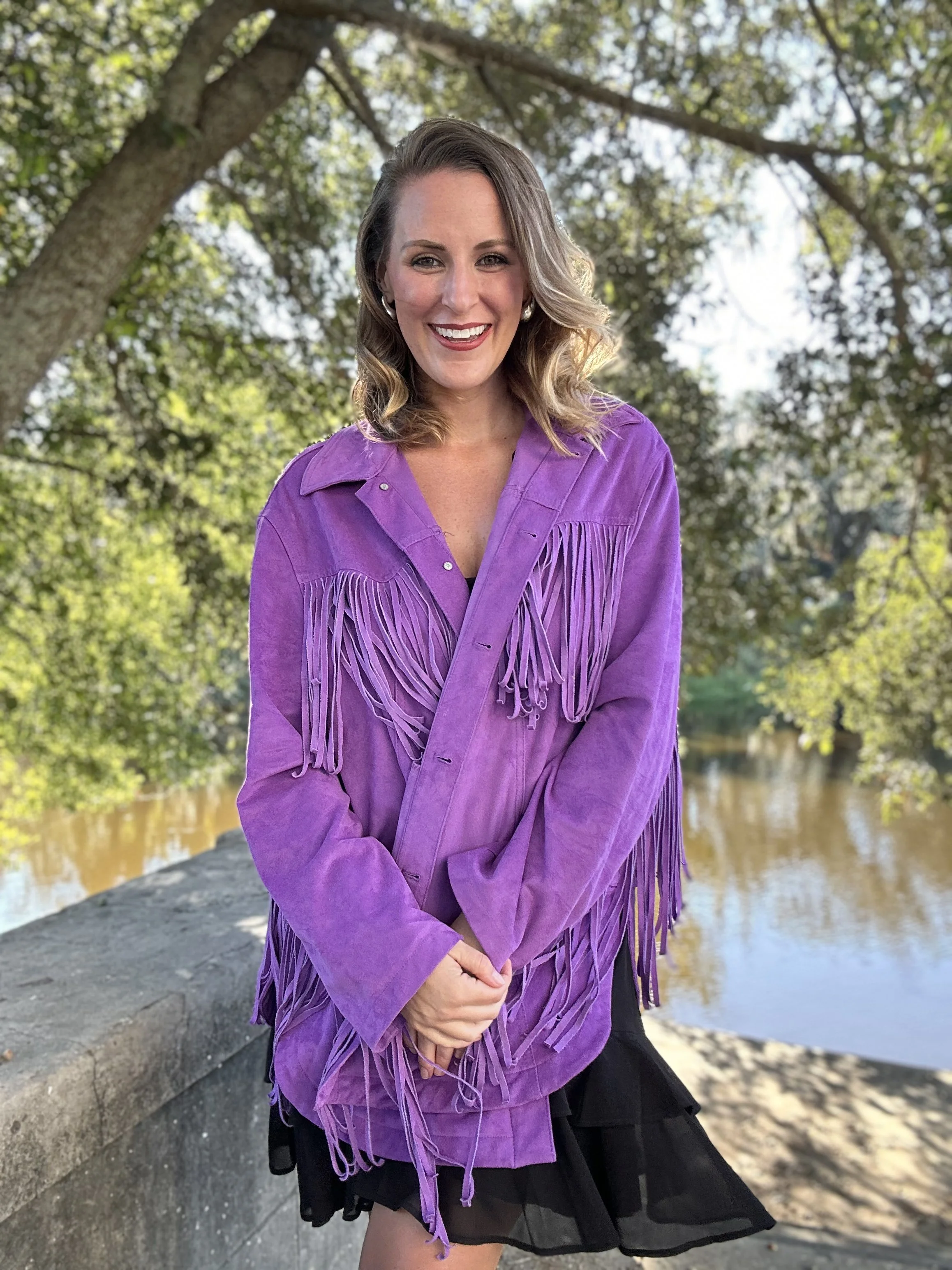 A woman with wavy blonde hair, smiling, stands outdoors near a body of water with trees in the background. She wears a purple fringed jacket and a black skirt.