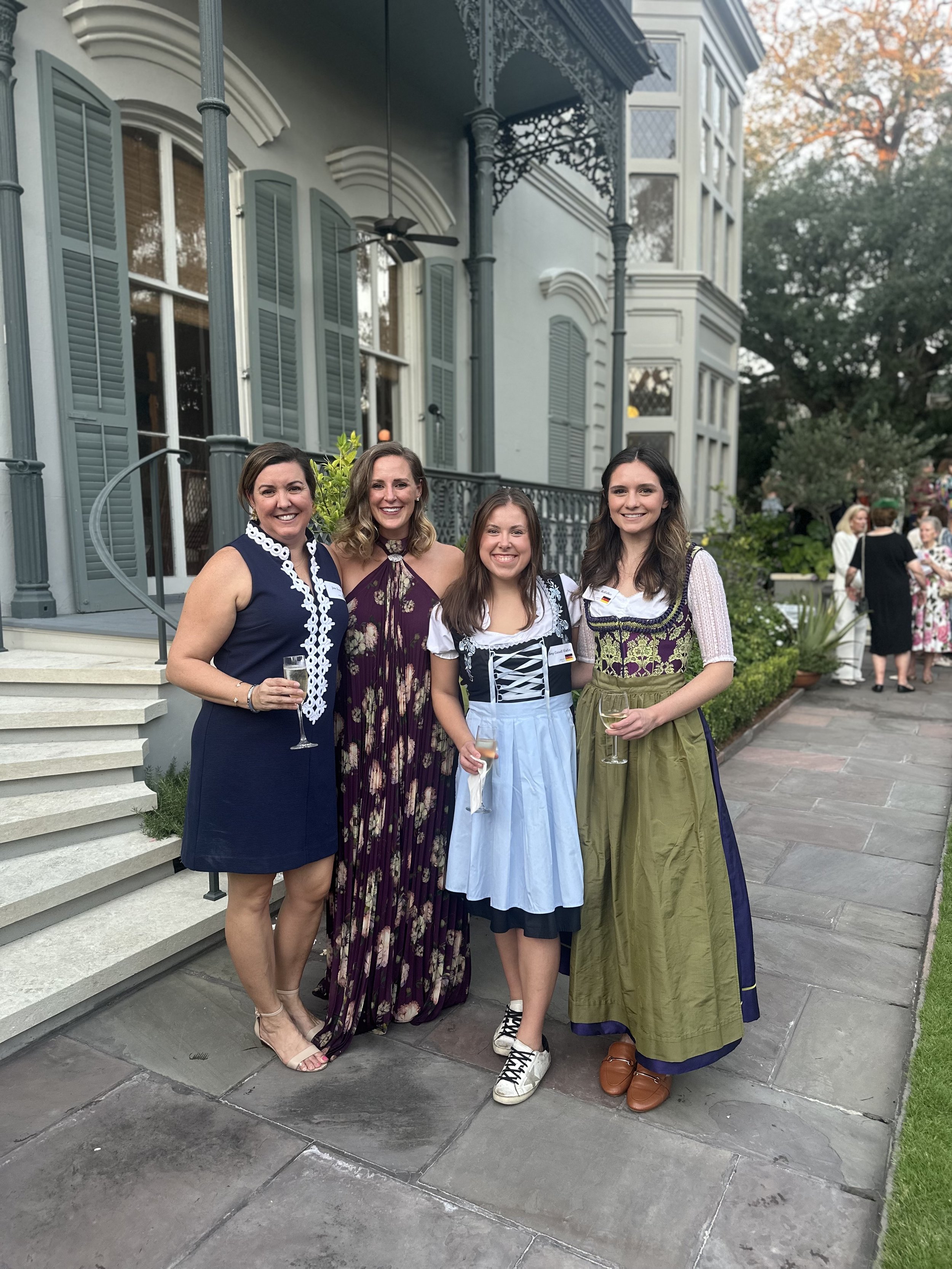 Four women standing outdoors in front of a large house, dressed in semi-formal attire, holding glasses of drinks and smiling at the camera.