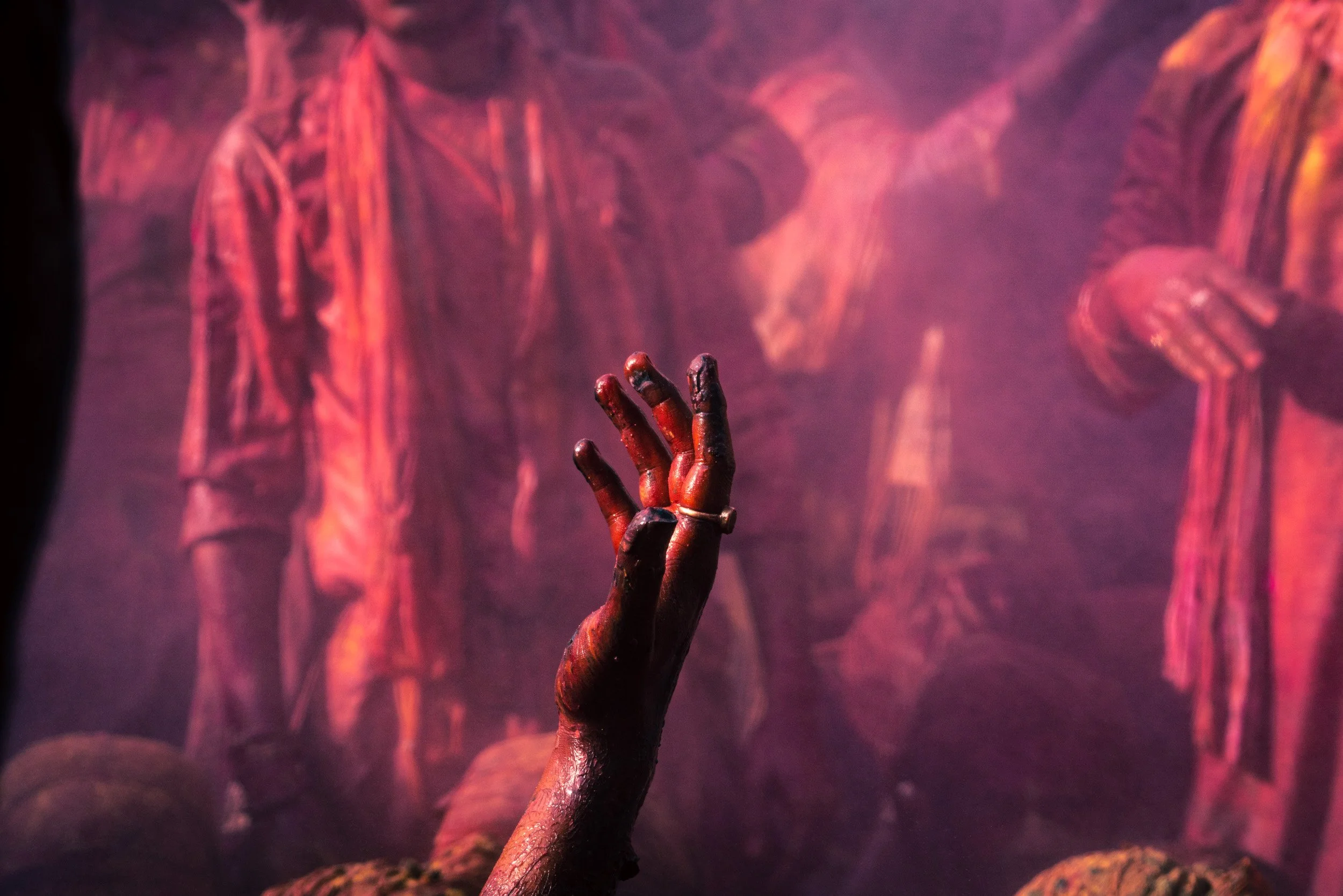 Close-up of a raised hand coated in red Holi powder reaching through a smoky cloud of pink gulal during a crowded Indian festival.