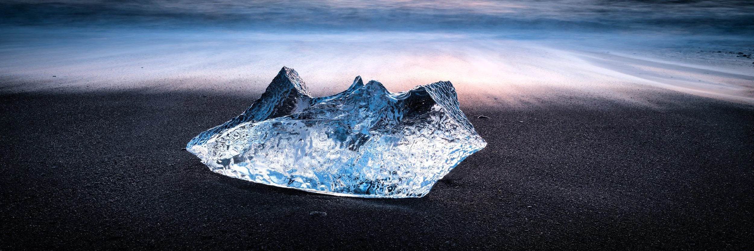 Blue iceberg on black volcanic sand at Diamond Beach in Iceland, with soft waves and pastel sunrise light.