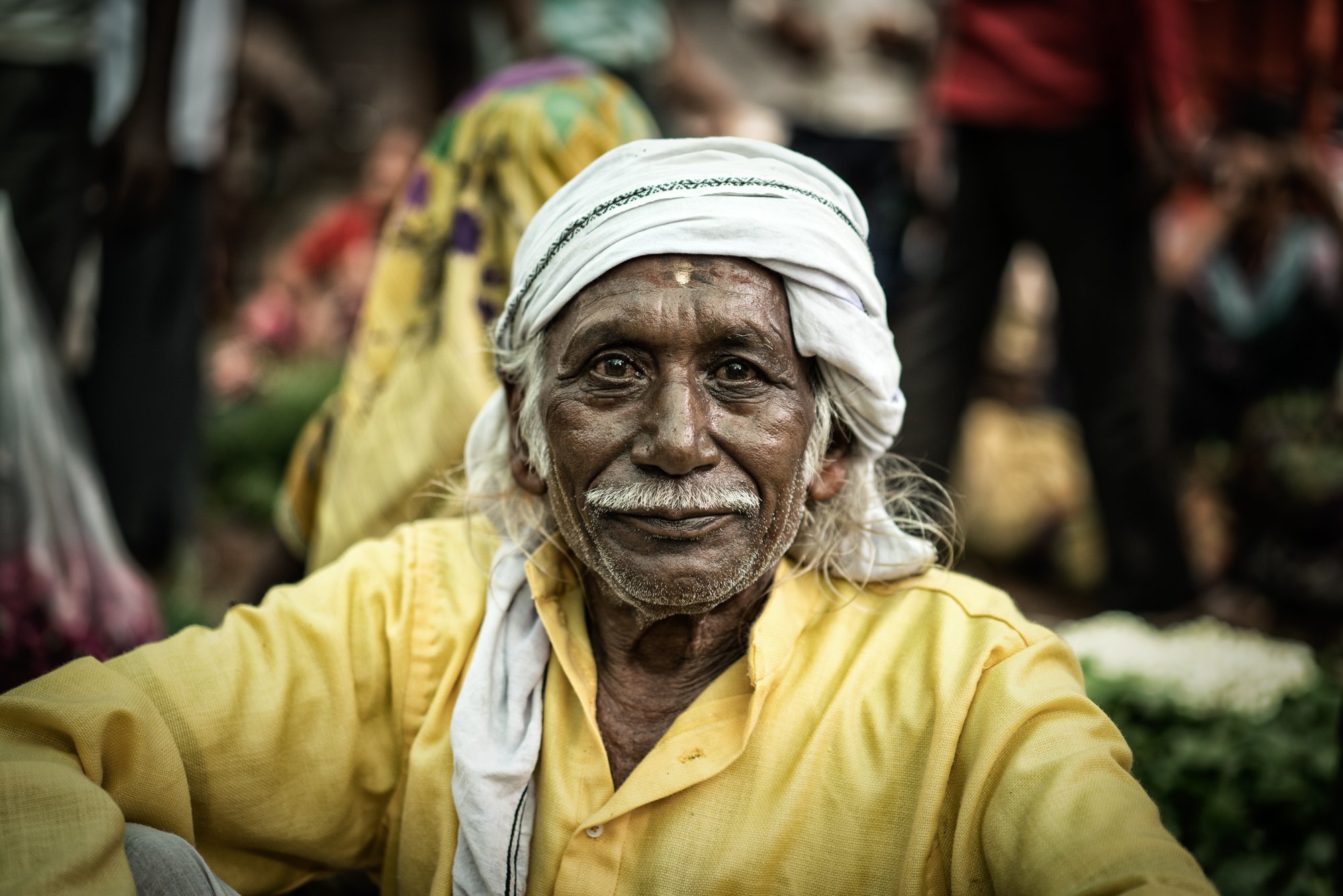 Elderly Indian man in a white turban and yellow shirt smiles for a close-up flower market portrait with blurred flowers in the background.