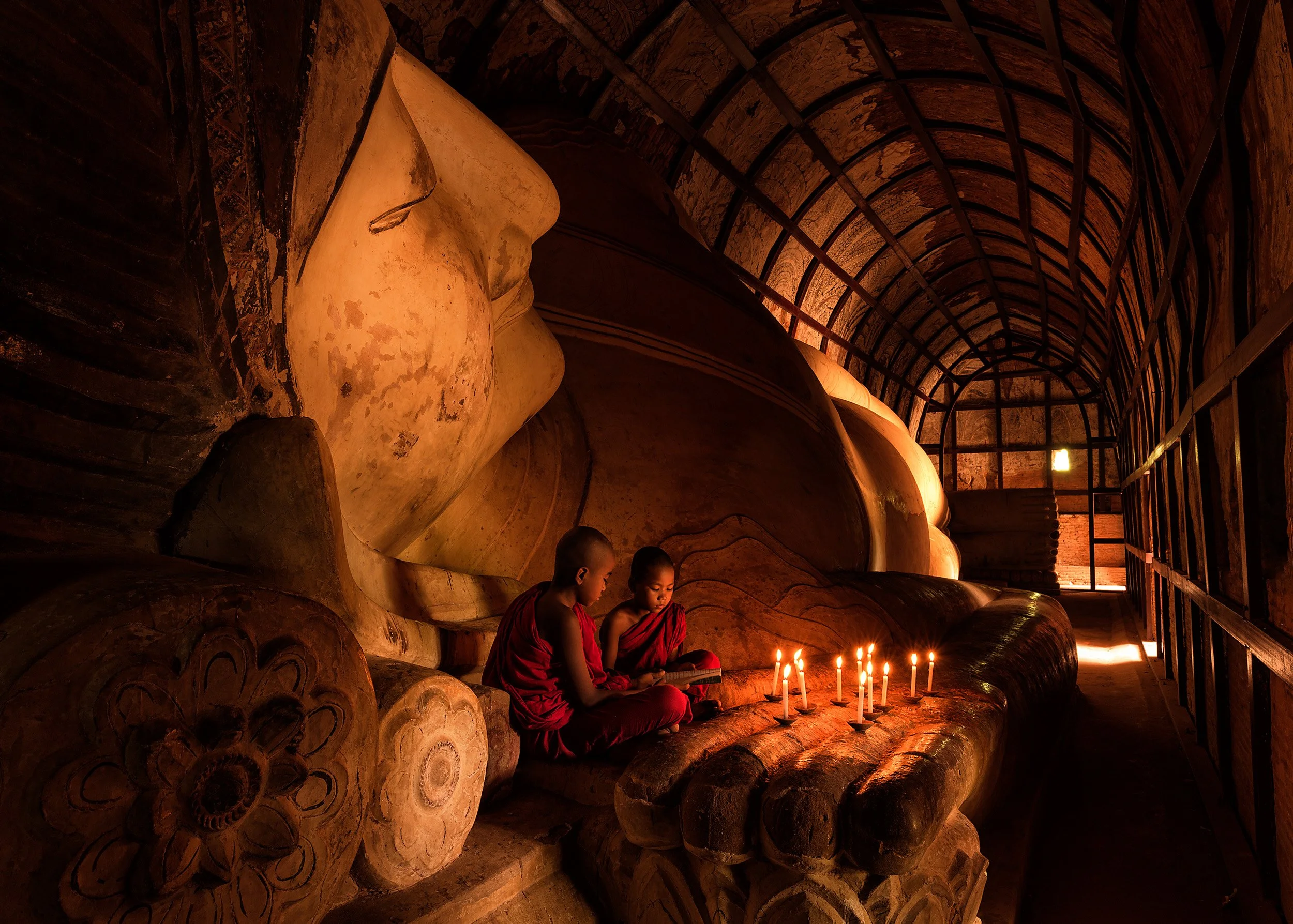 Young buddhist monks lighting candles beside a reclining Buddha inside a warmly lit Myanmar temple.
