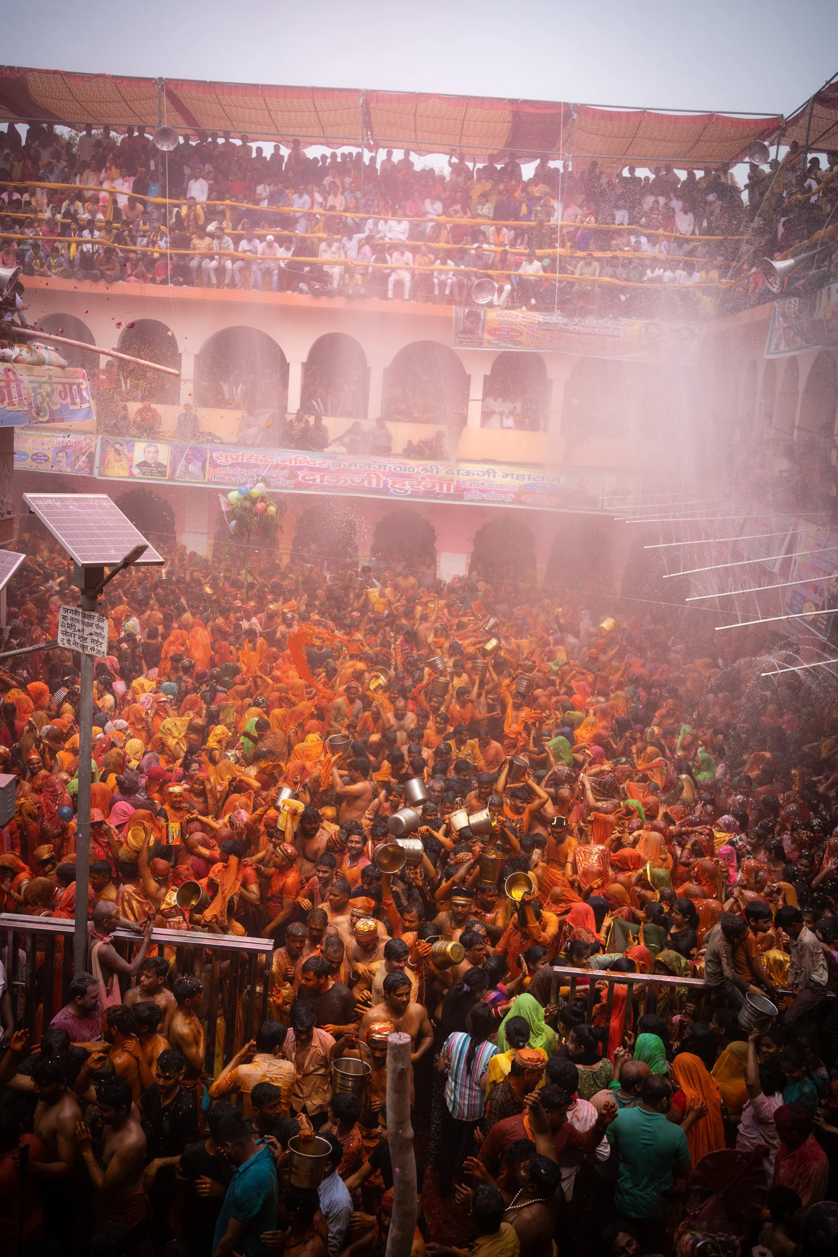 Wide view of a packed Indian Holi festival courtyard, where devotees throw water and colored powder beneath balconies, banners, and misty pink gulal.