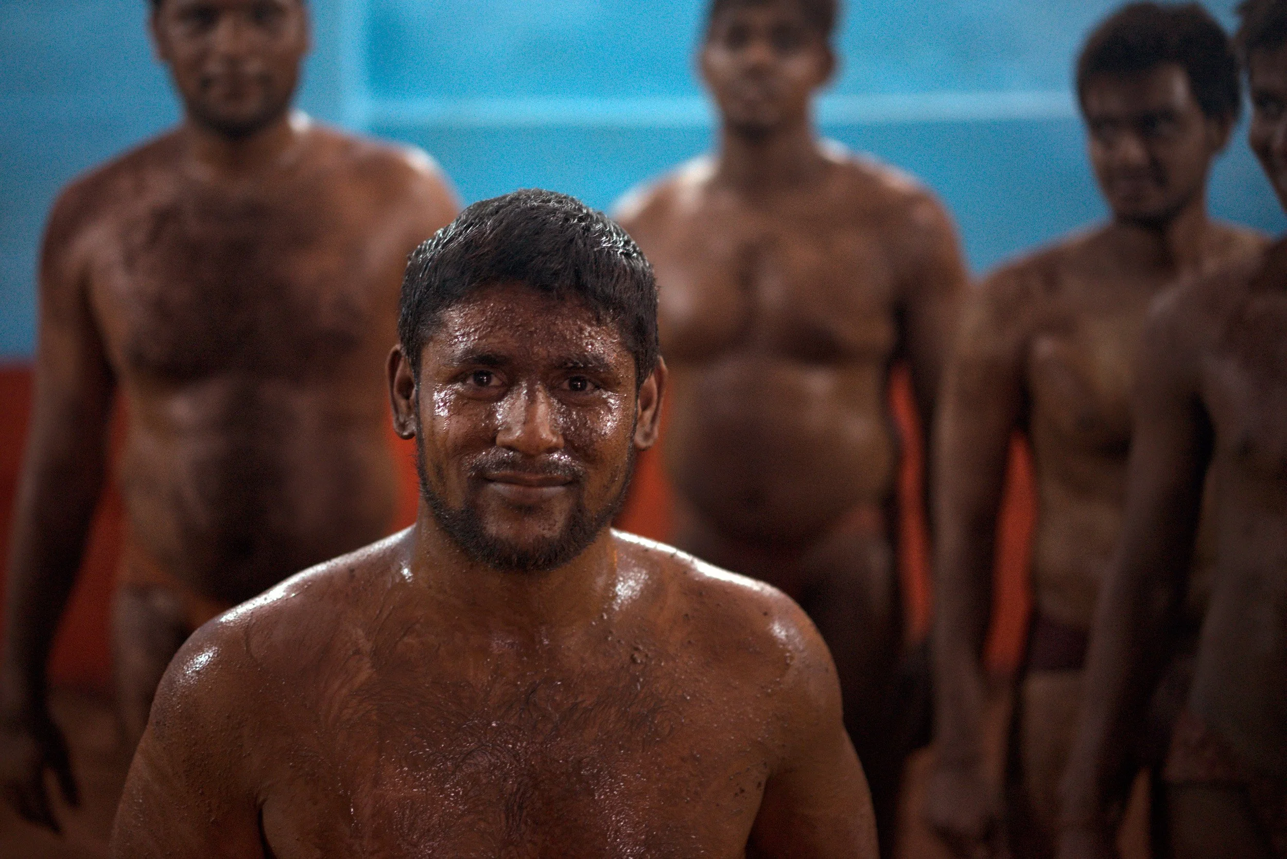 Traditional Indian kushti wrestlers stand in a mud-floor akhara, with shirtless athletes in orange loincloths against blue and red walls.