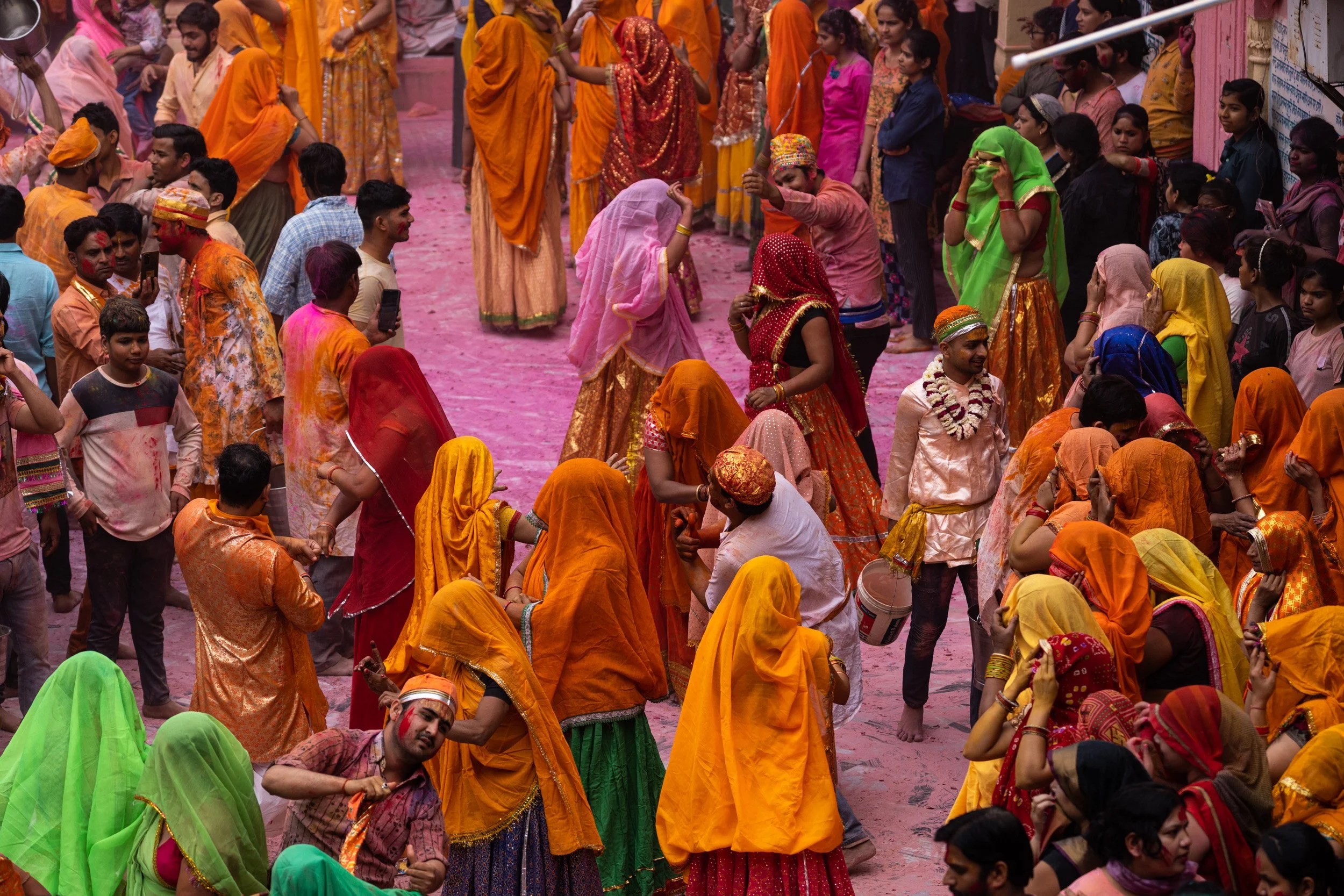 Large Holi crowd in bright yellow, orange, red, and green traditional clothing gathers in a temple courtyard filled with pink powder stains.
