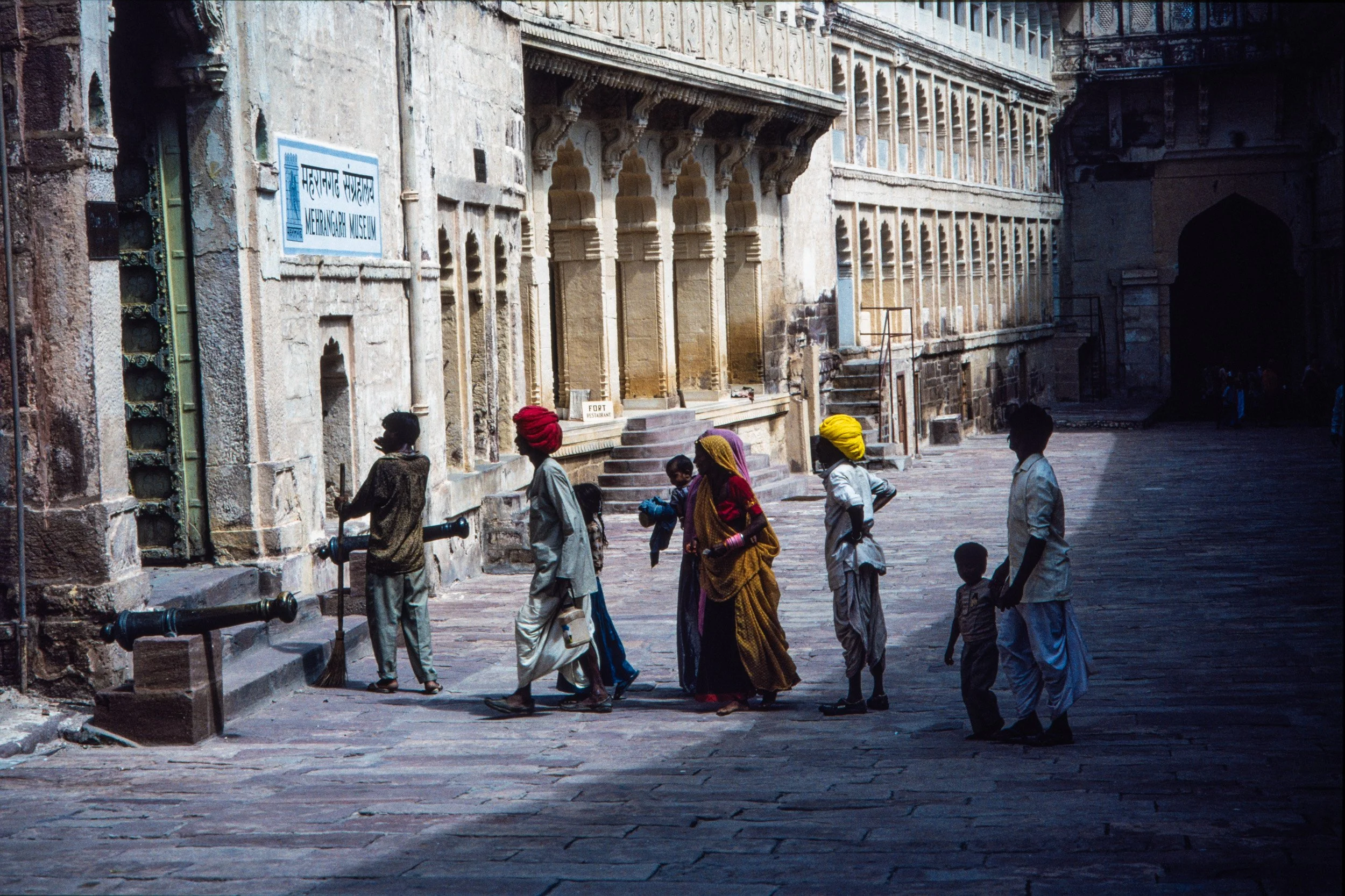 Visitors walk through the stone courtyard of Mehrangarh Museum, with historic fort architecture, cannons, arched windows, and colorful turbans in Jodhpur, India.