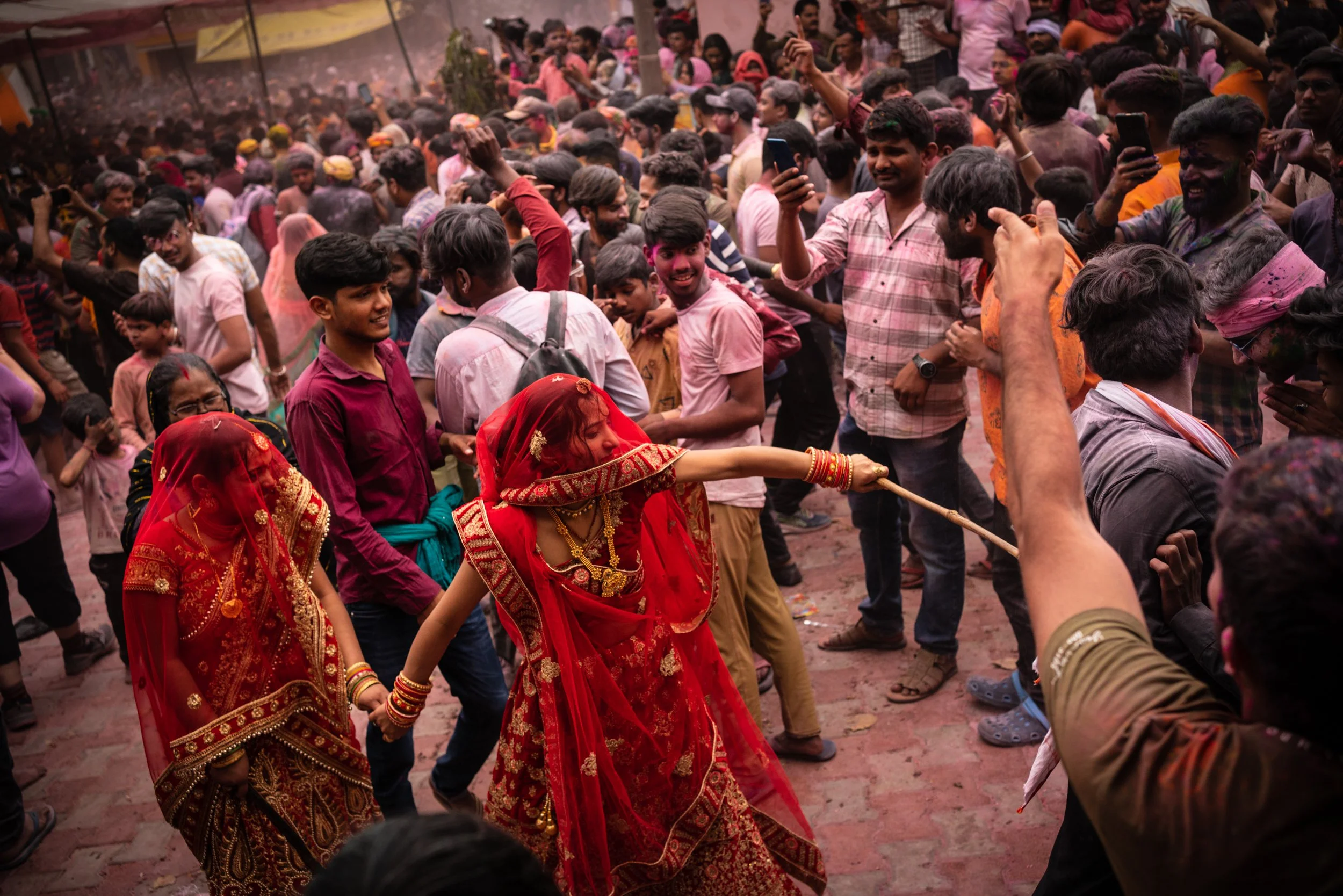 Women in red bridal-style veils play Lathmar Holi with sticks in a crowded Indian temple courtyard, surrounded by festivalgoers covered in pink gulal powder.