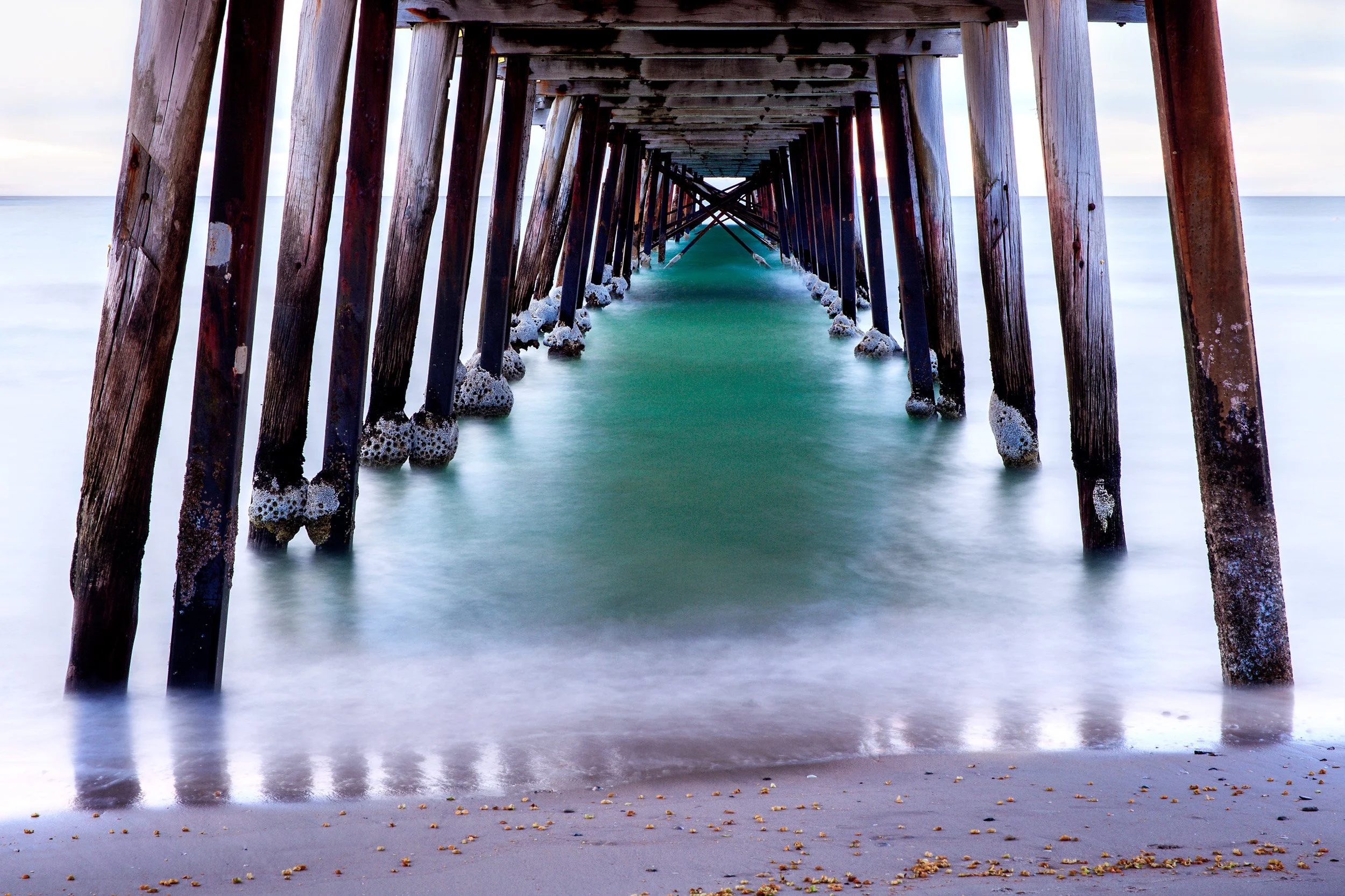 Long exposure view beneath Henley Beach Jetty in South Australia, with timber pylons framing smooth turquoise water.