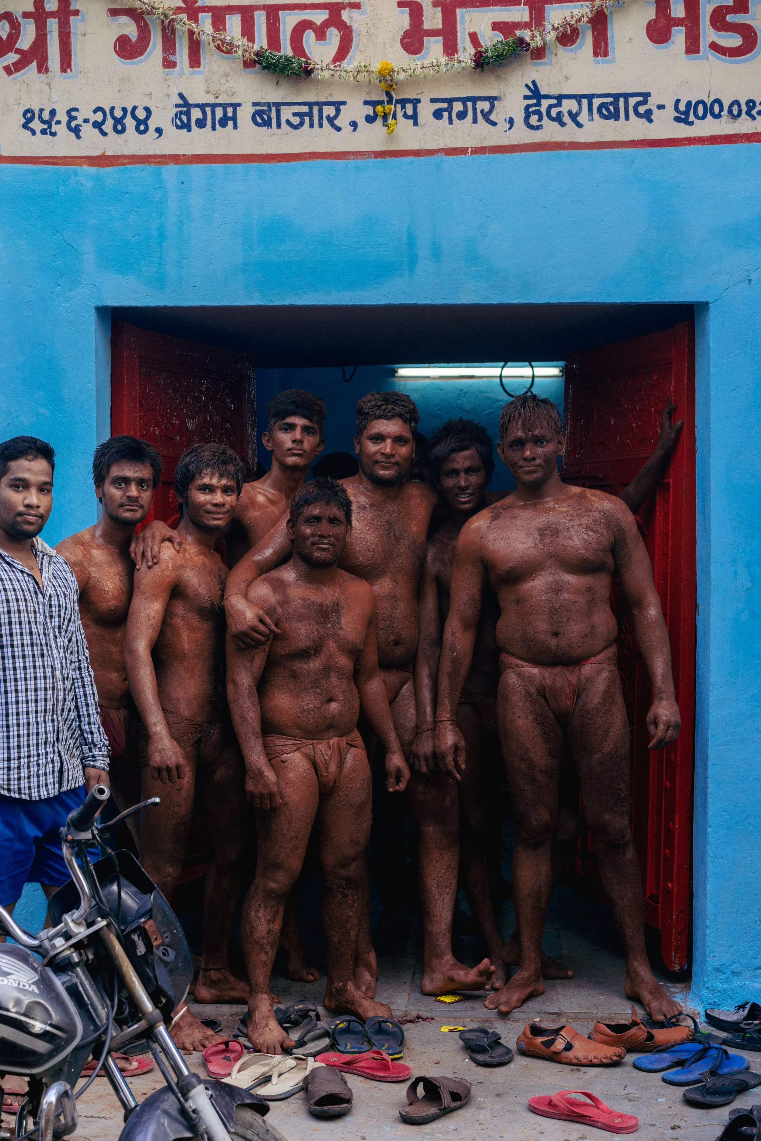 Group portrait of Indian kushti wrestlers covered in red clay outside a blue akhara entrance, with sandals, a motorbike, and Hindi signage showing “बेगम बाजार” and “हैदराबाद.”
